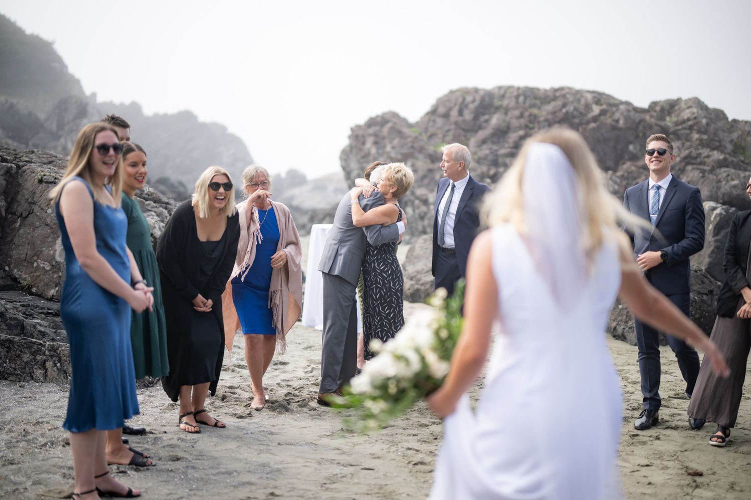 Wedding party gathers on dramatic rocky beach for celebration.