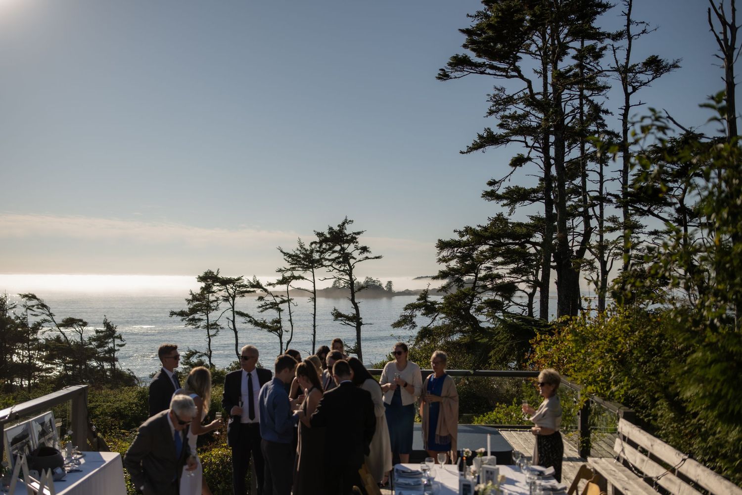 Wedding guests gather on wooden deck overlooking Pacific Ocean coastline.