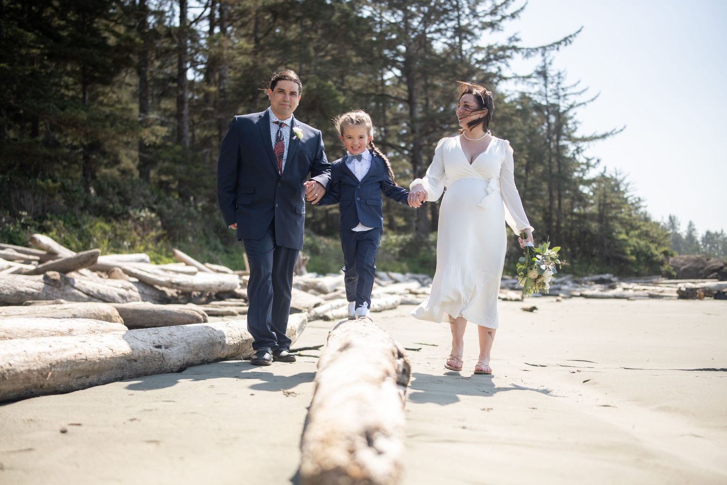 Wedding party poses on beach with driftwood and evergreen trees.
