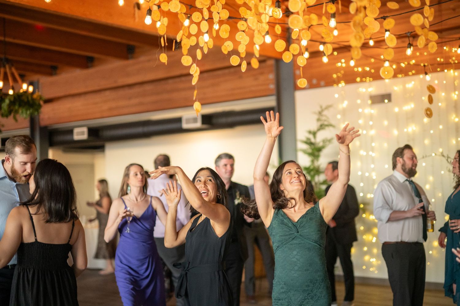 Guests dancing and celebrating at an indoor wedding reception decorated with hanging lights and wooden beams.