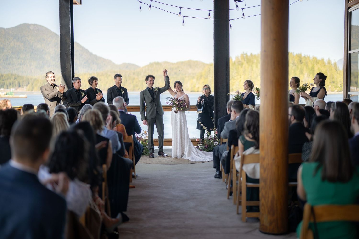 Wedding guests mingle during cocktail hour at a covered outdoor venue with string lights and wooden beams.