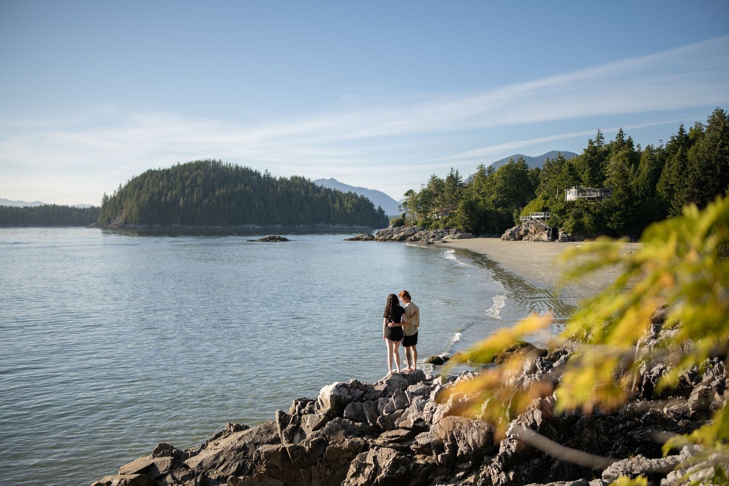 Couple embraces on rocky coastal cliffs overlooking a serene bay with mountains in the background.