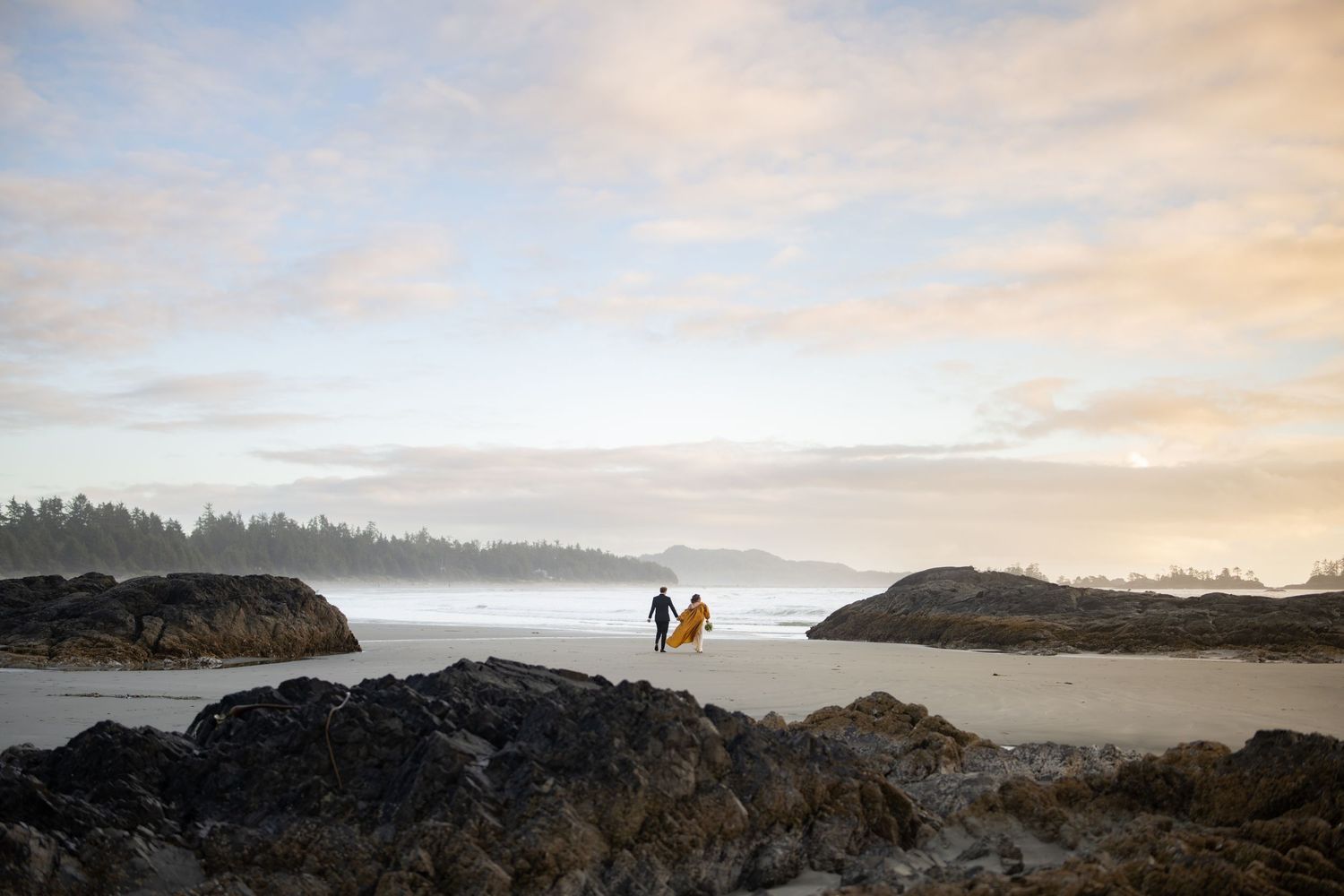 Silhouettes of people walking between rocky formations on a foggy beach at dusk.