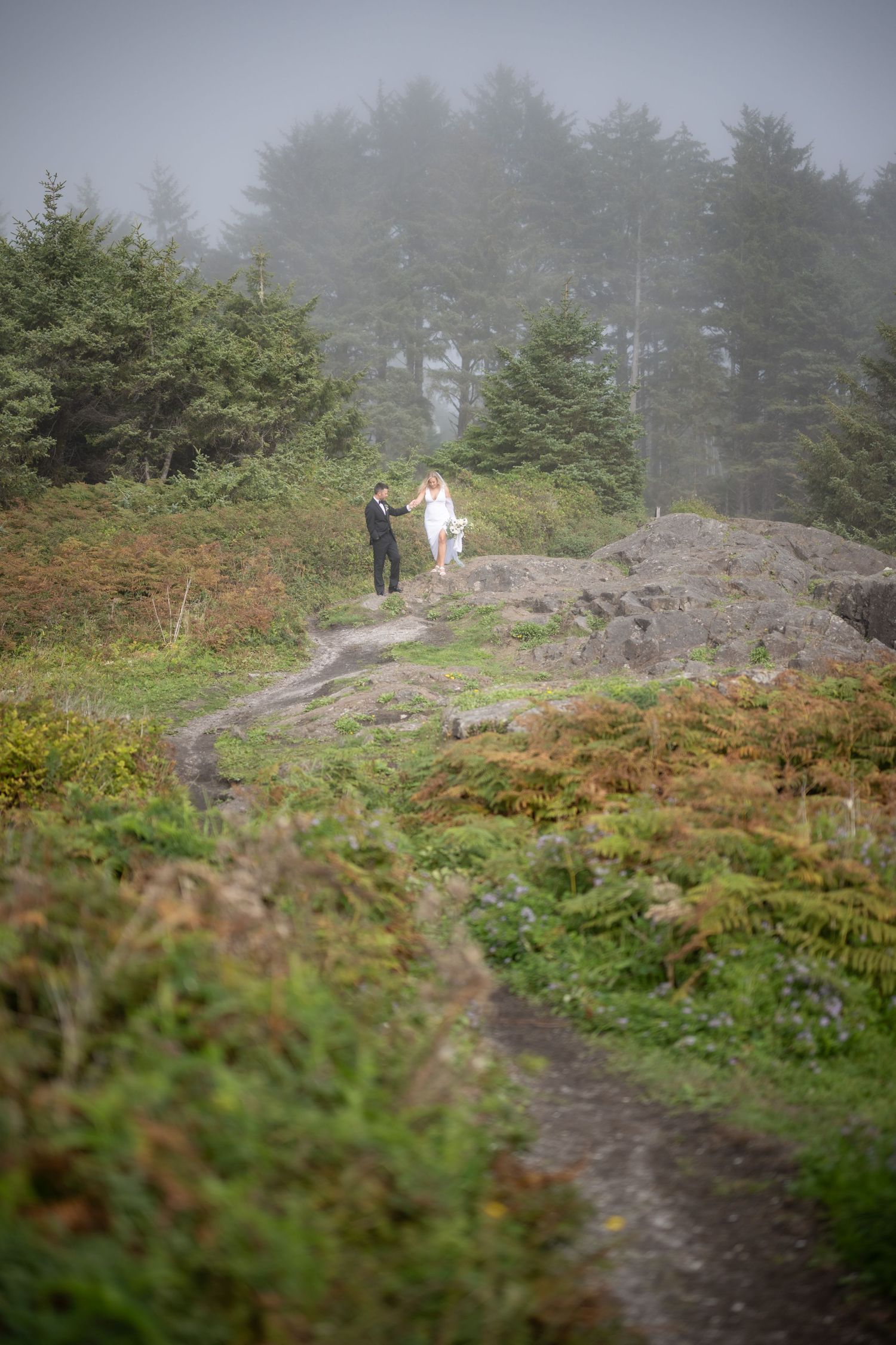 A couple walking along a misty mountain trail surrounded by evergreen trees and ferns.