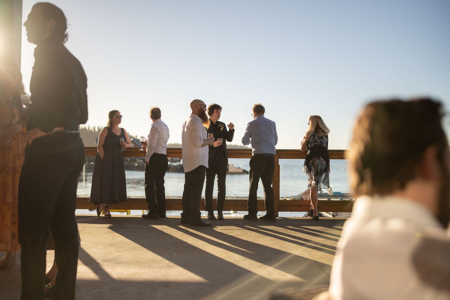 Silhouettes of wedding guests gathered on a deck during golden hour.