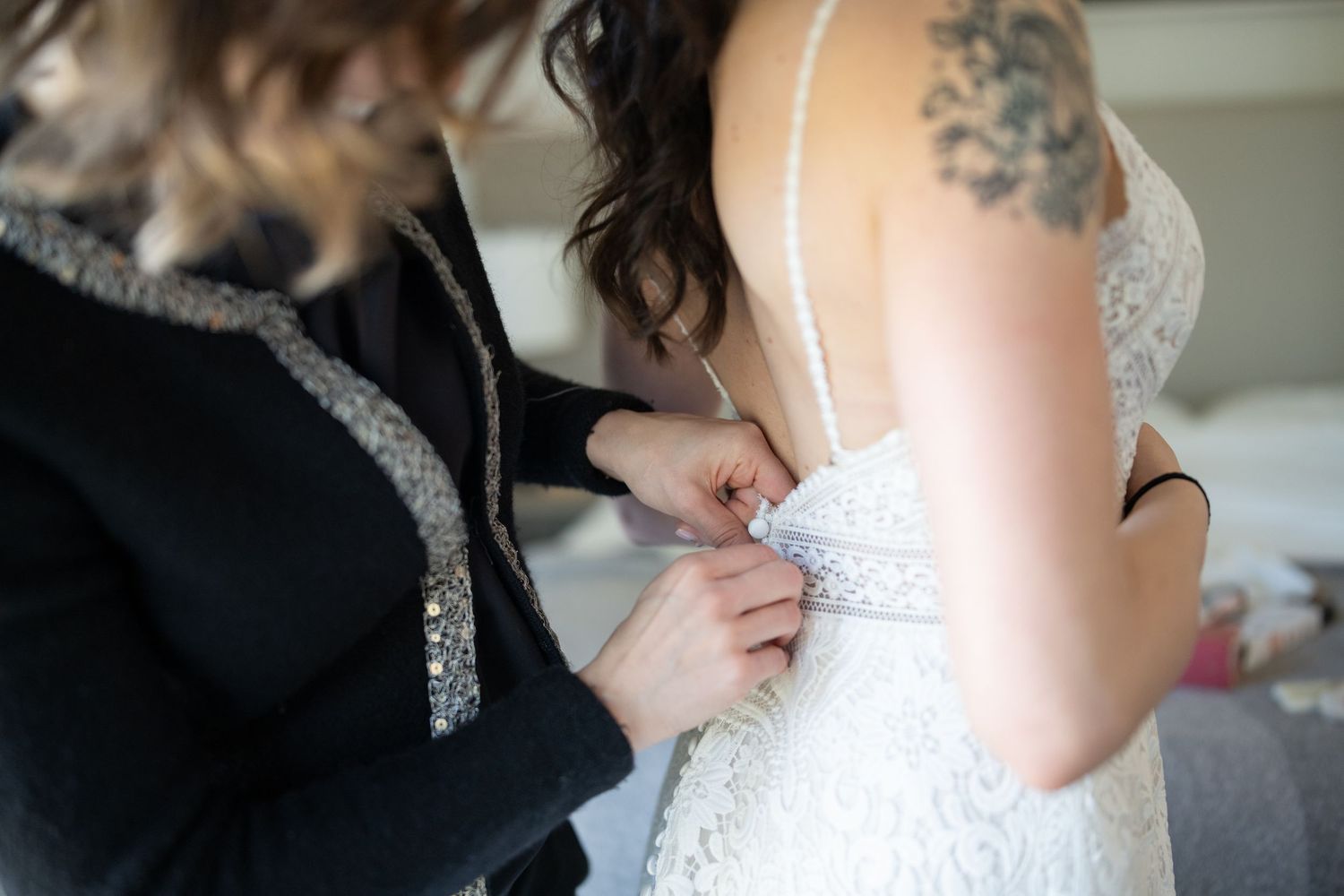 A close-up shot of someone helping to fasten a white lace wedding dress with visible tattoo detail.