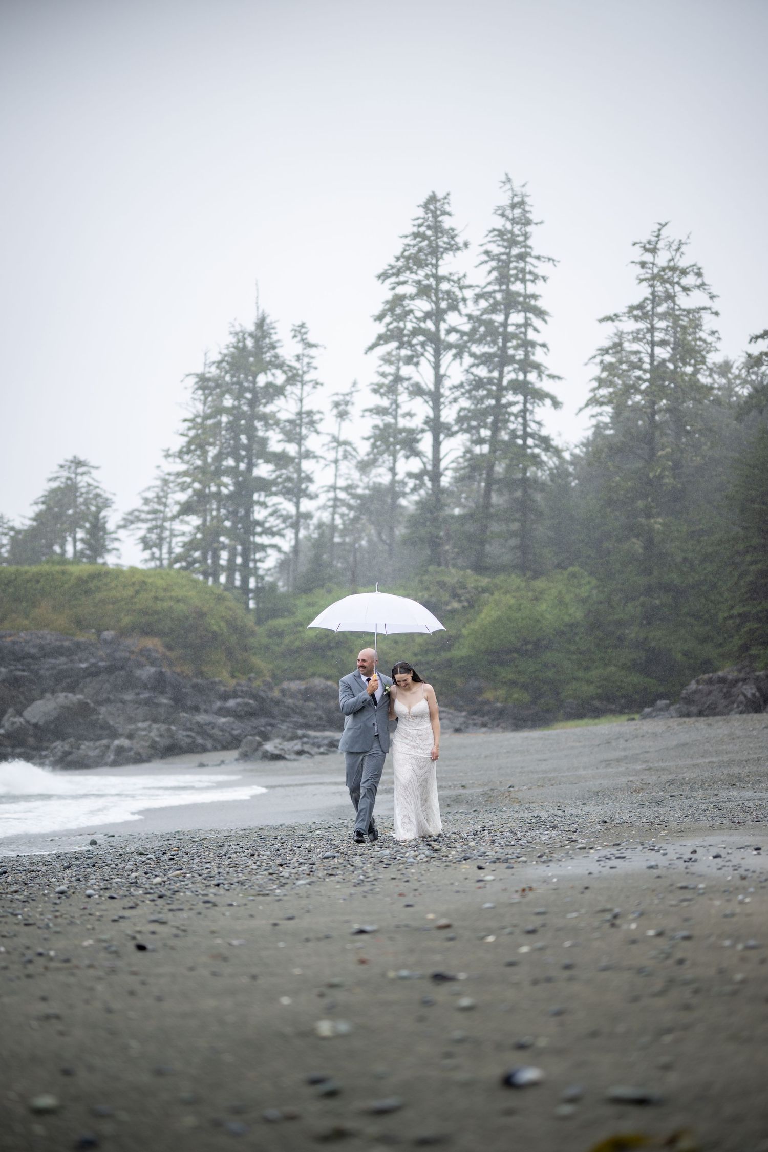 A couple stands together with an umbrella on a foggy beach with tall evergreen trees in the background.