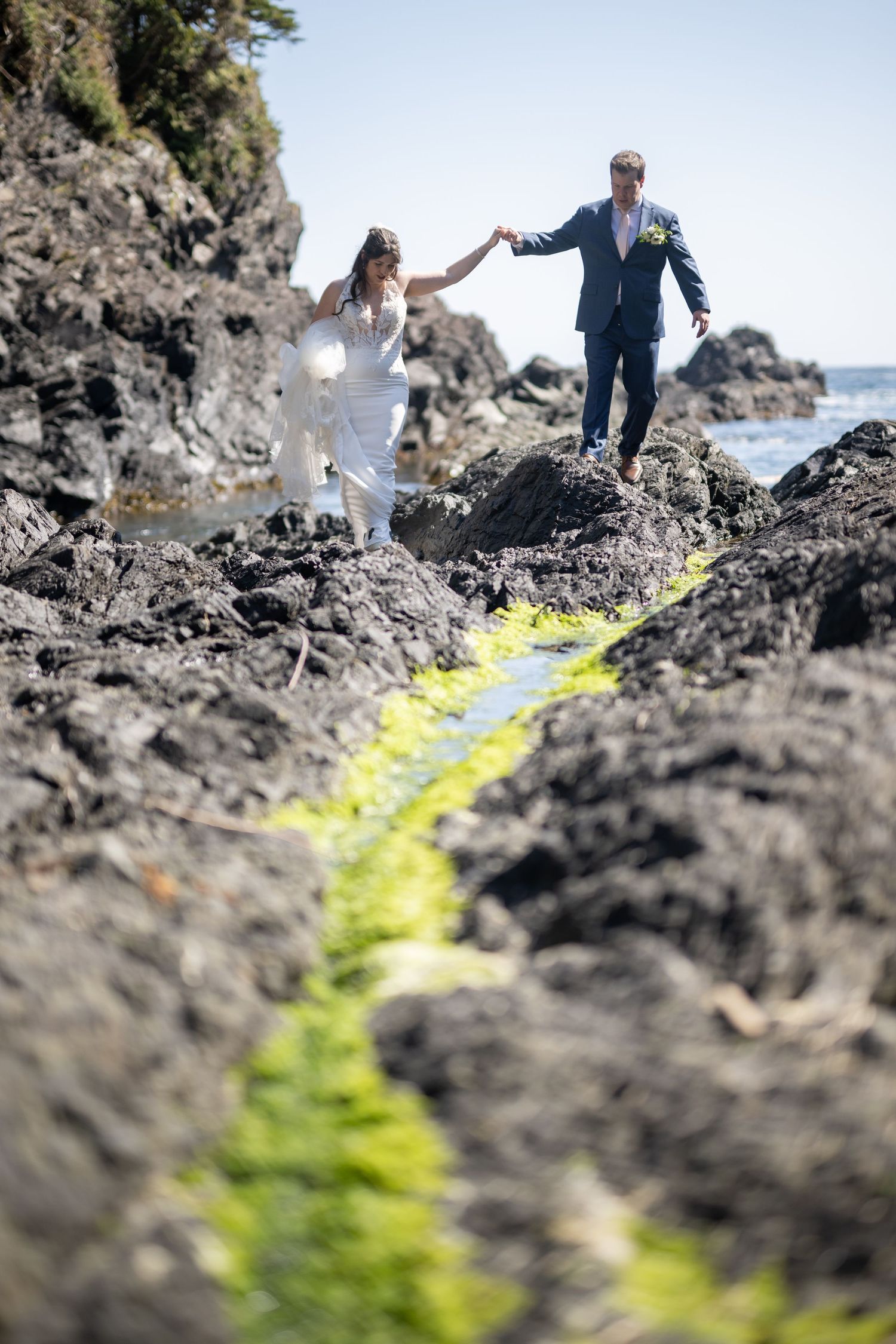 Wedding ceremony participants walking along mossy coastal rocks by the ocean.