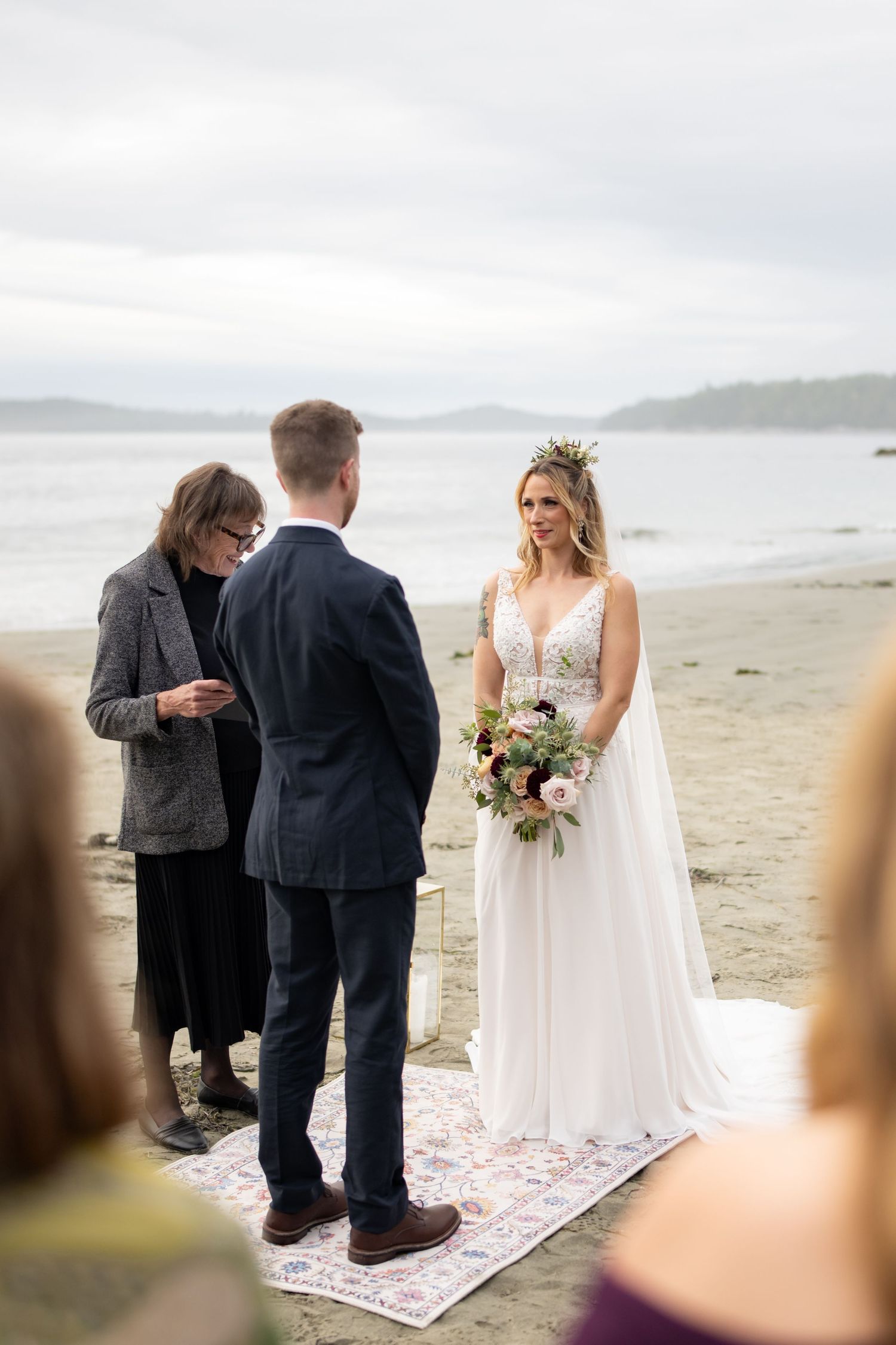 An intimate beach wedding ceremony takes place on a decorative rug with overcast skies.
