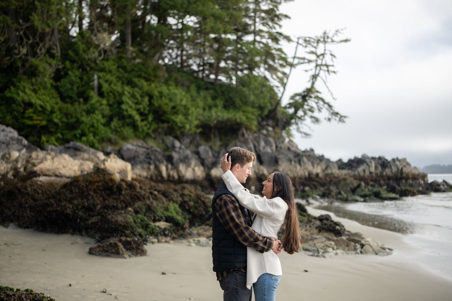 Romantic moment captured on rocky beach with evergreen trees in background.