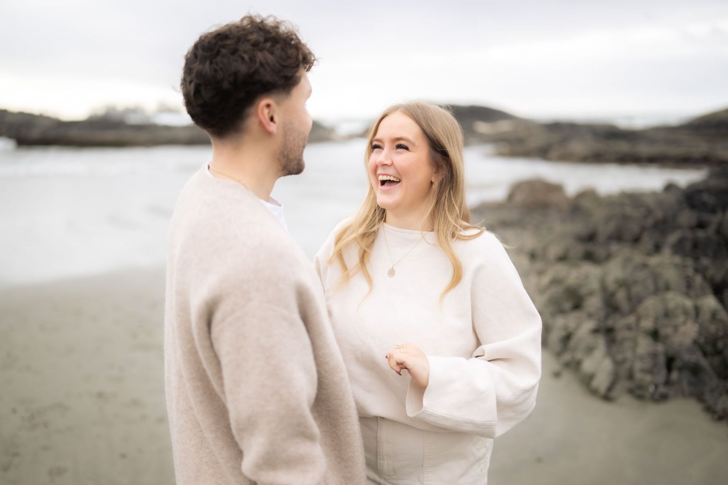 Happy couple laughing together on rocky beach during engagement photoshoot.