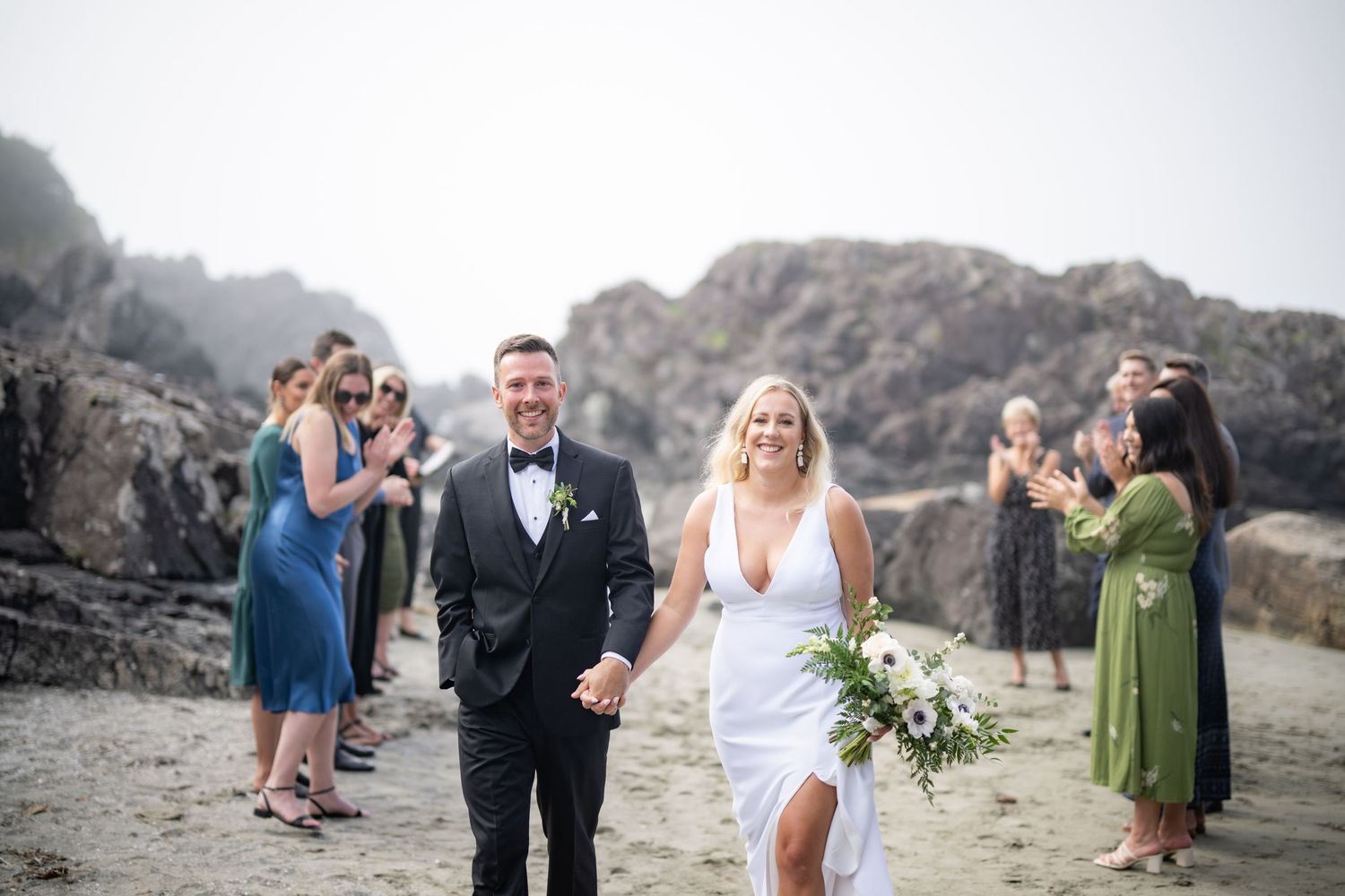 Newlyweds walk joyfully on a beach path while wedding guests celebrate behind them.