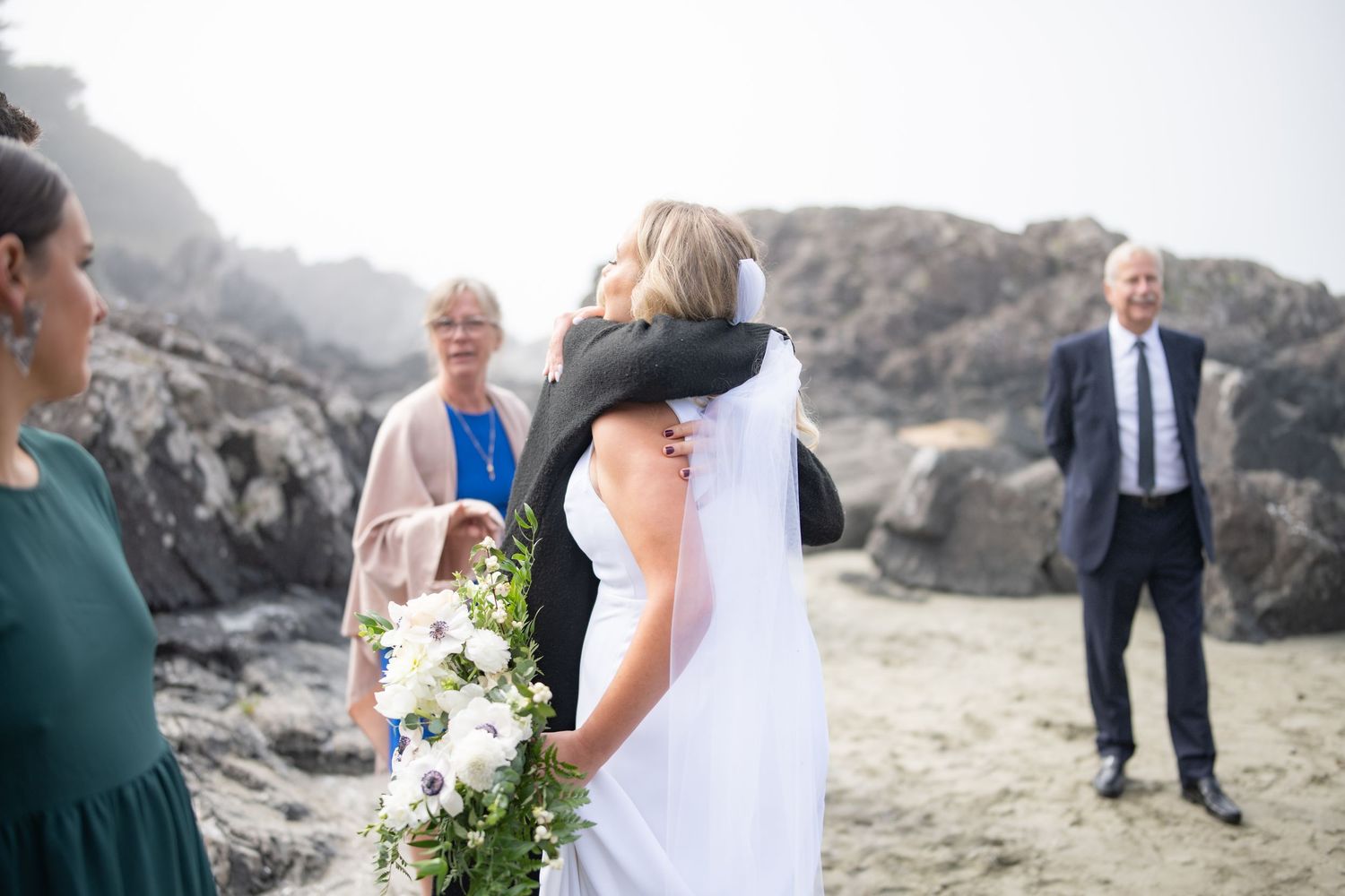 Wedding guests mingle on sandy beach with dramatic rocky coastline in background.