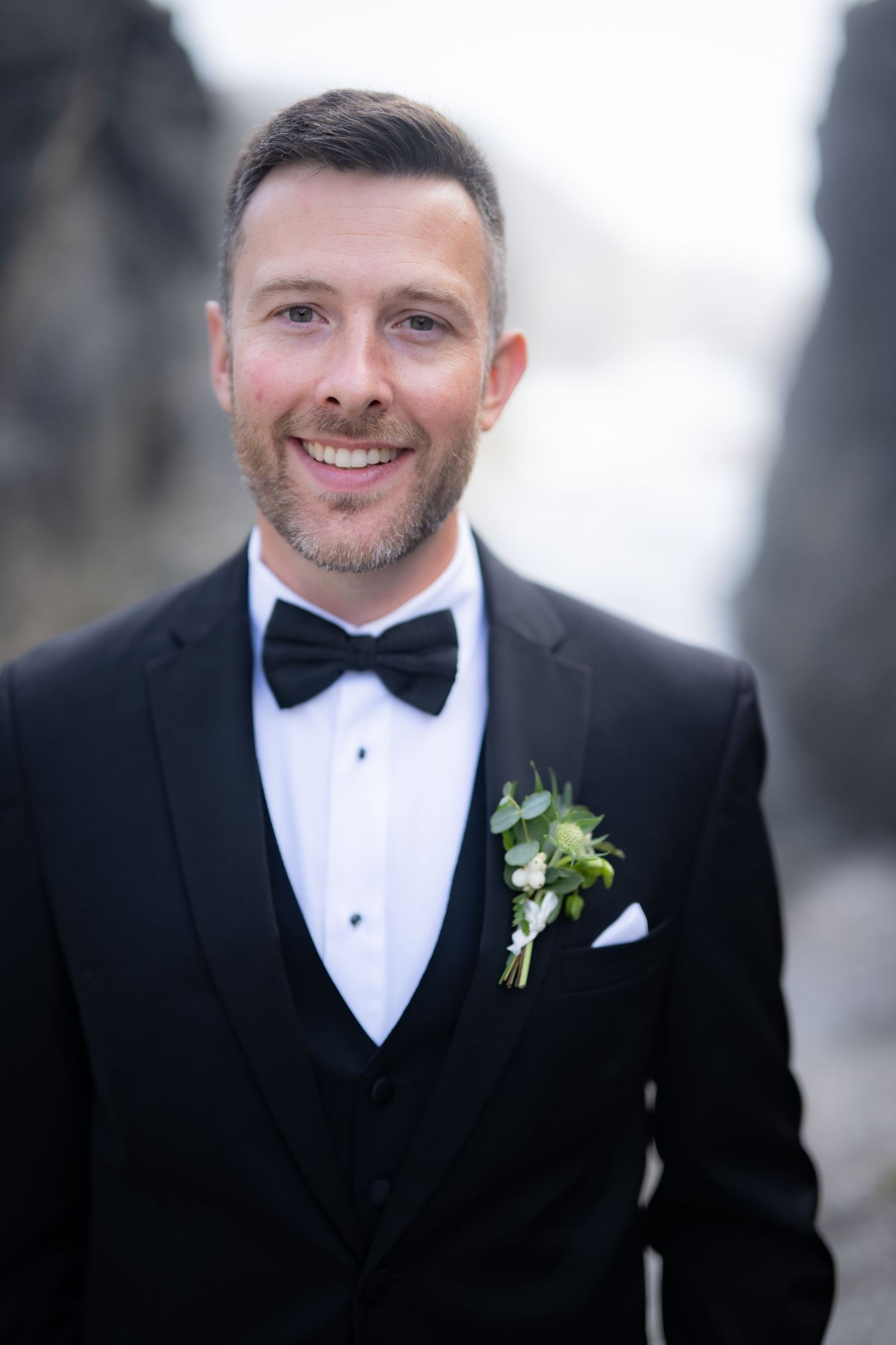 Groom in classic black tuxedo and bow tie smiles during wedding photoshoot.