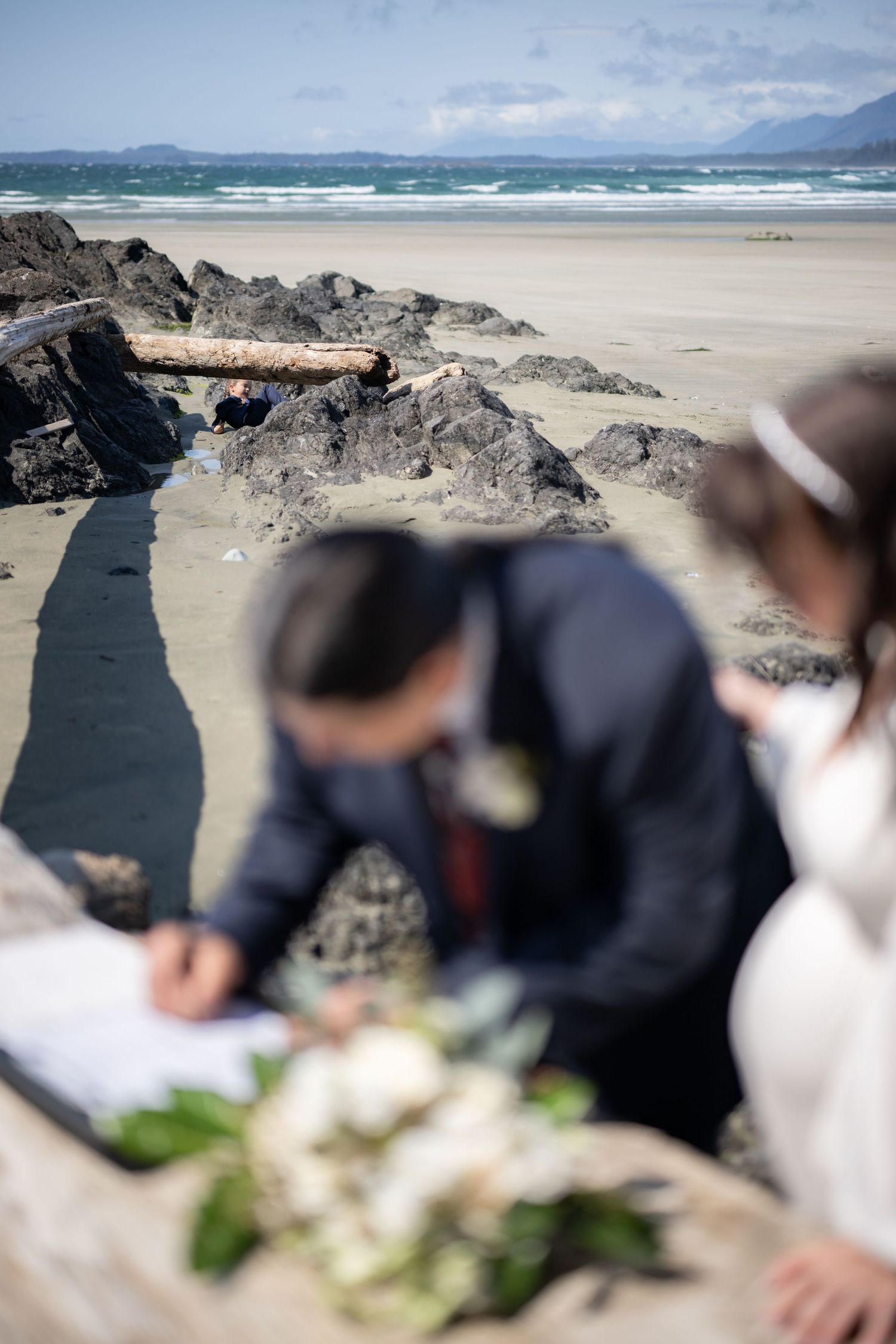 Wedding party gathers on scenic rocky beach with waves and mountains in background.