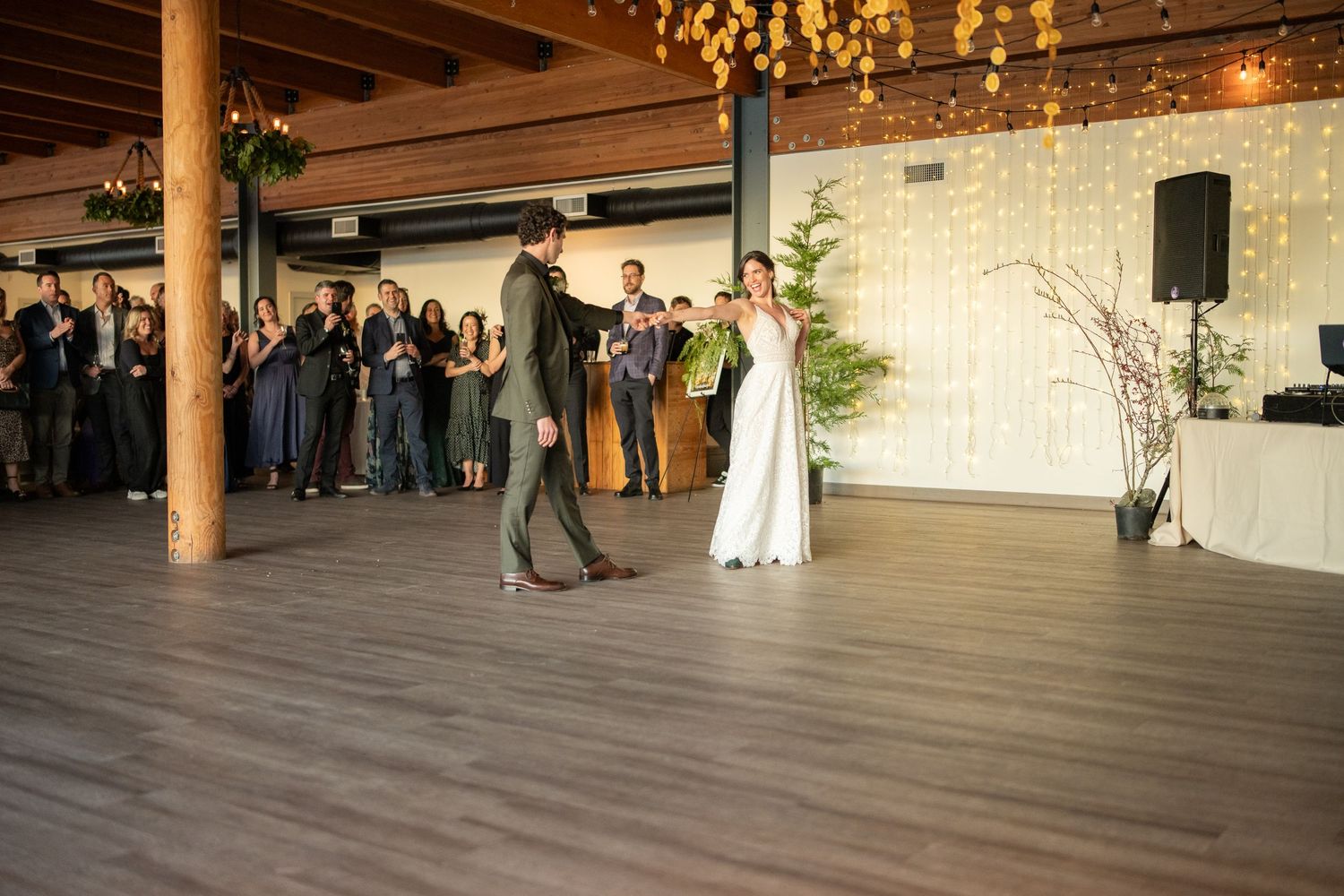 Wedding guests watching couple's first dance in spacious reception hall with string lights.