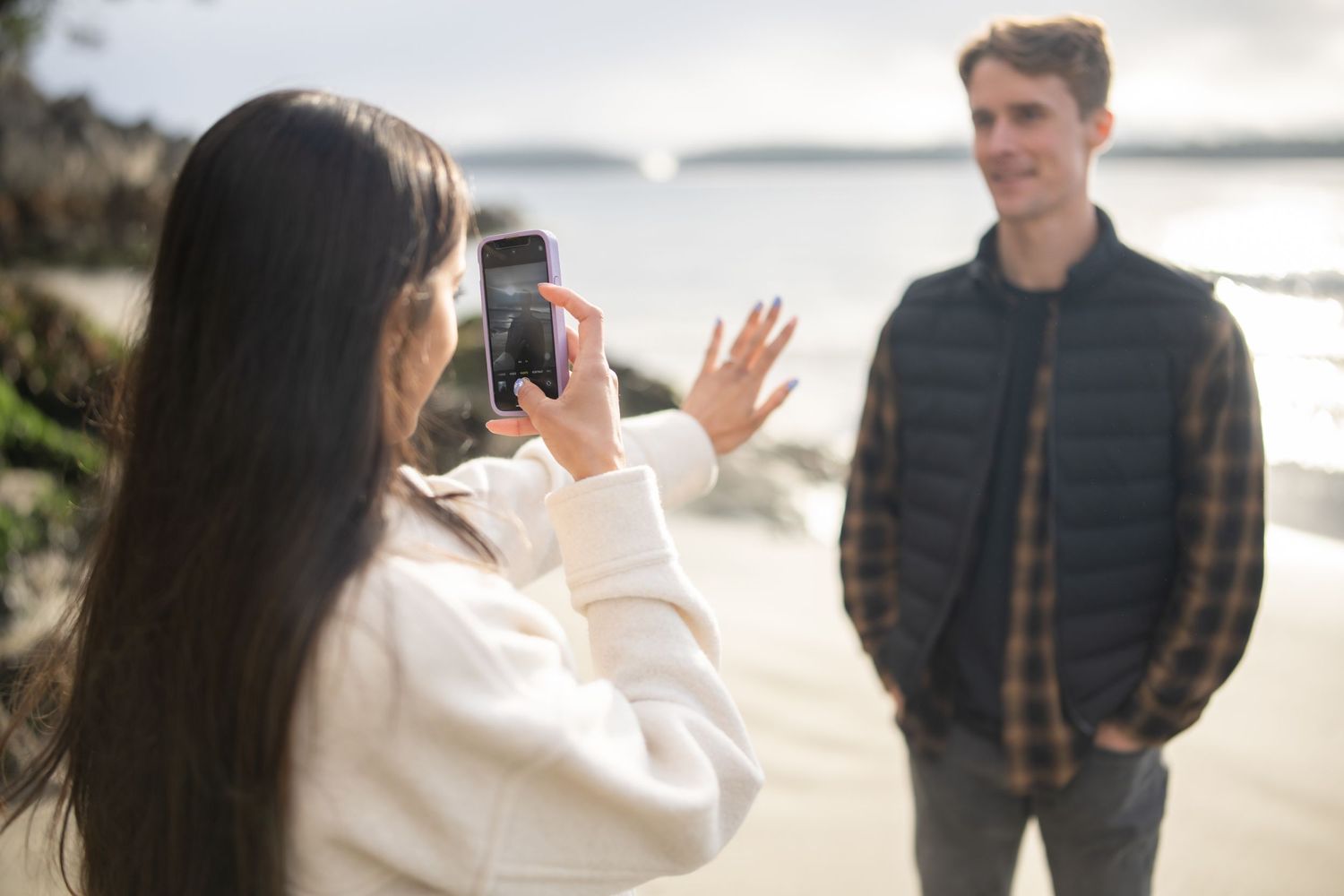 Person in white sweater taking photo with phone on scenic beach.