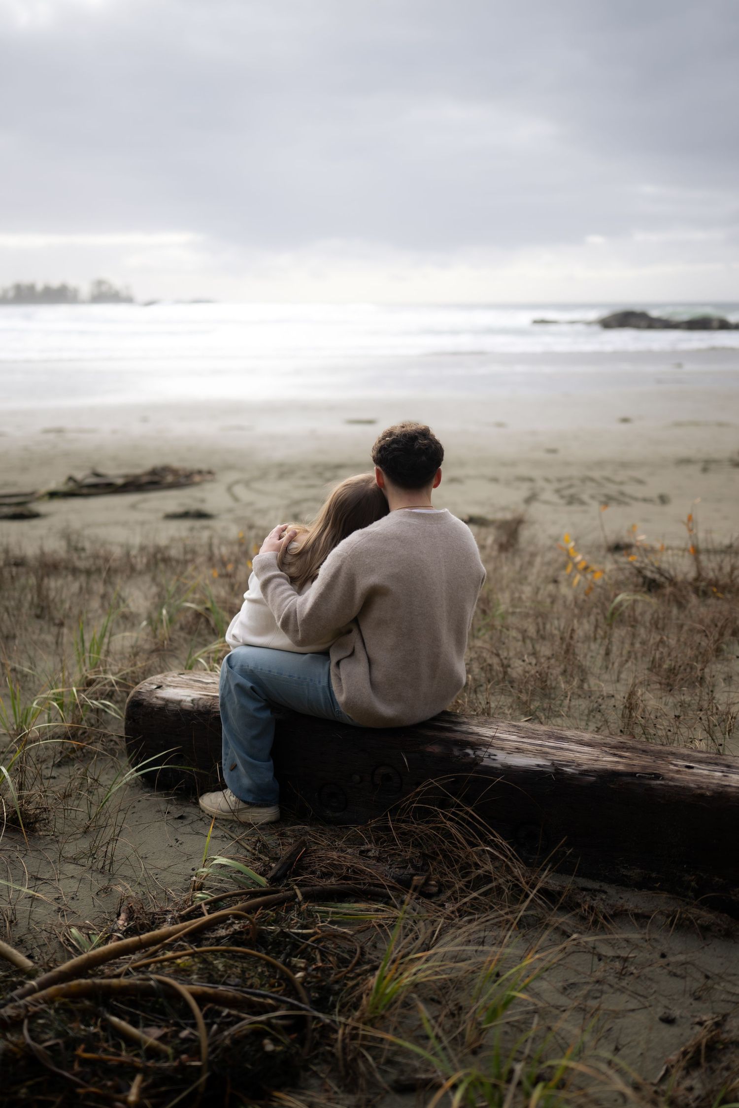 Moody coastal landscape with couple resting on driftwood surrounded by beach grass.
