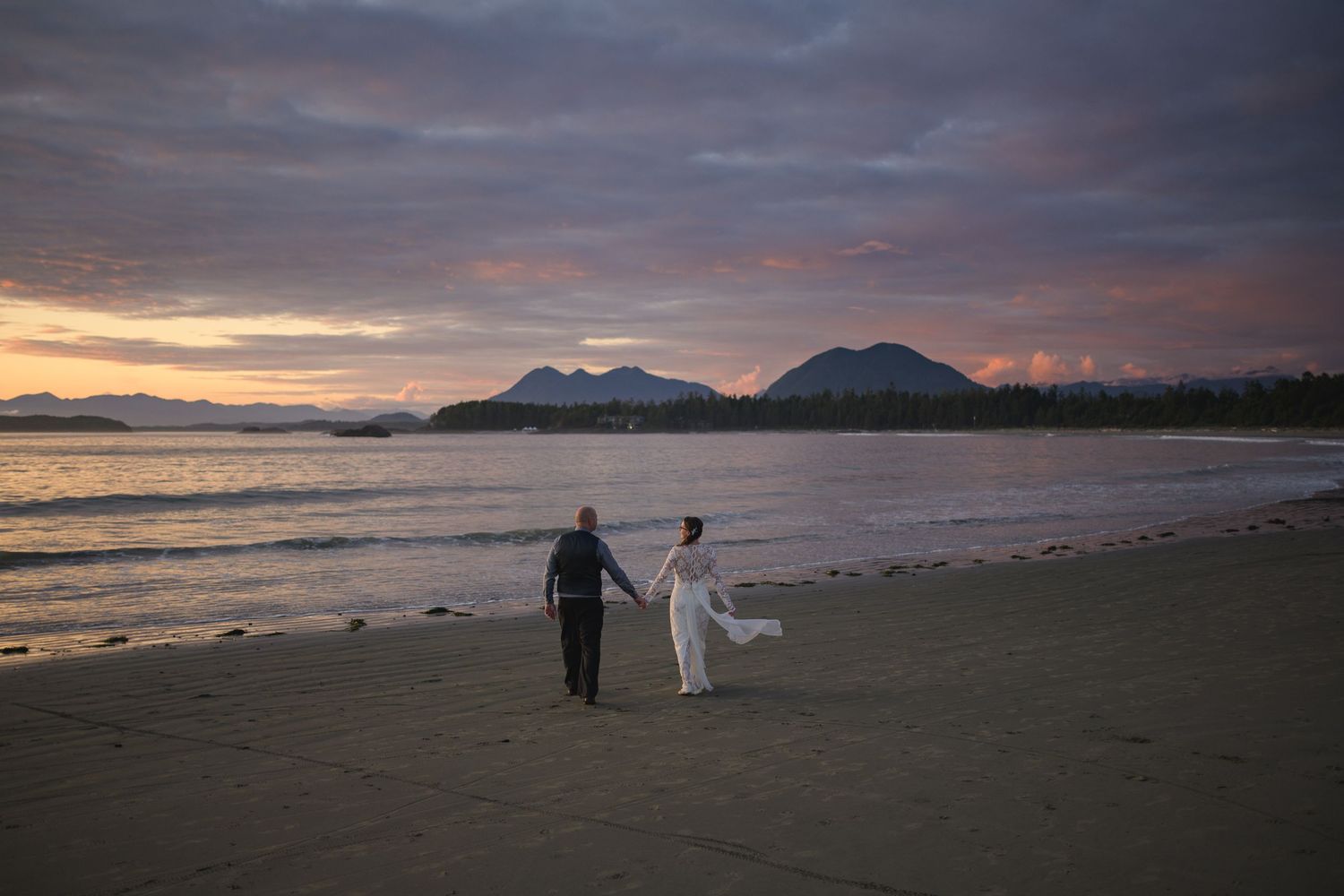 A couple walks along a beach at sunset with mountains visible in the distance.