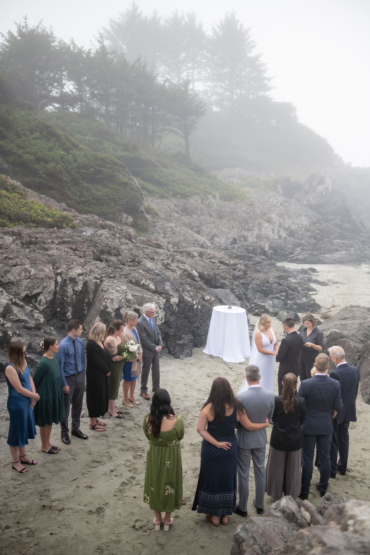 An intimate wedding ceremony gathering on a foggy Pacific Northwest beach with rocky cliffs.