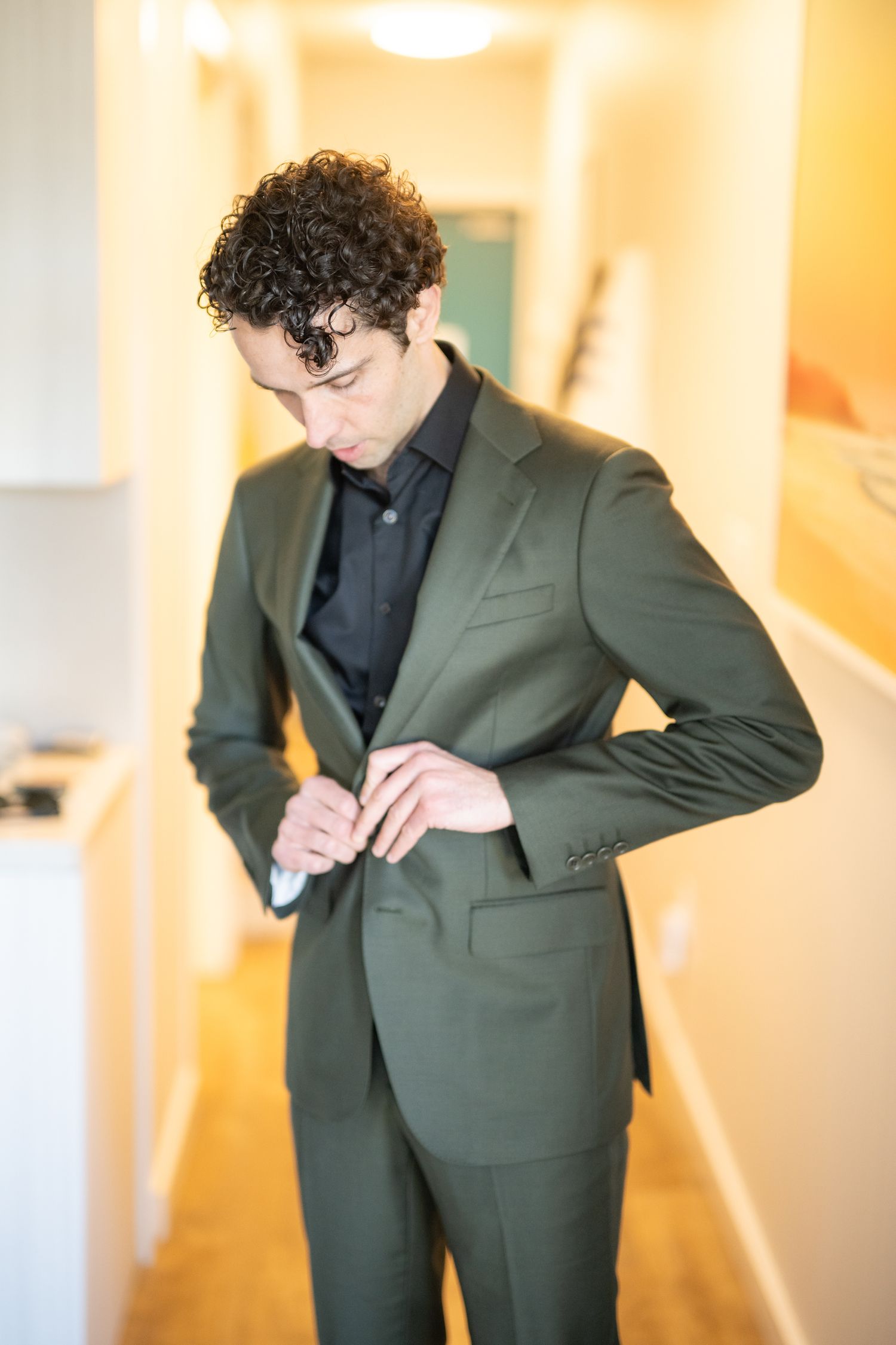 Person adjusting the buttons on an olive green suit jacket in a hallway.