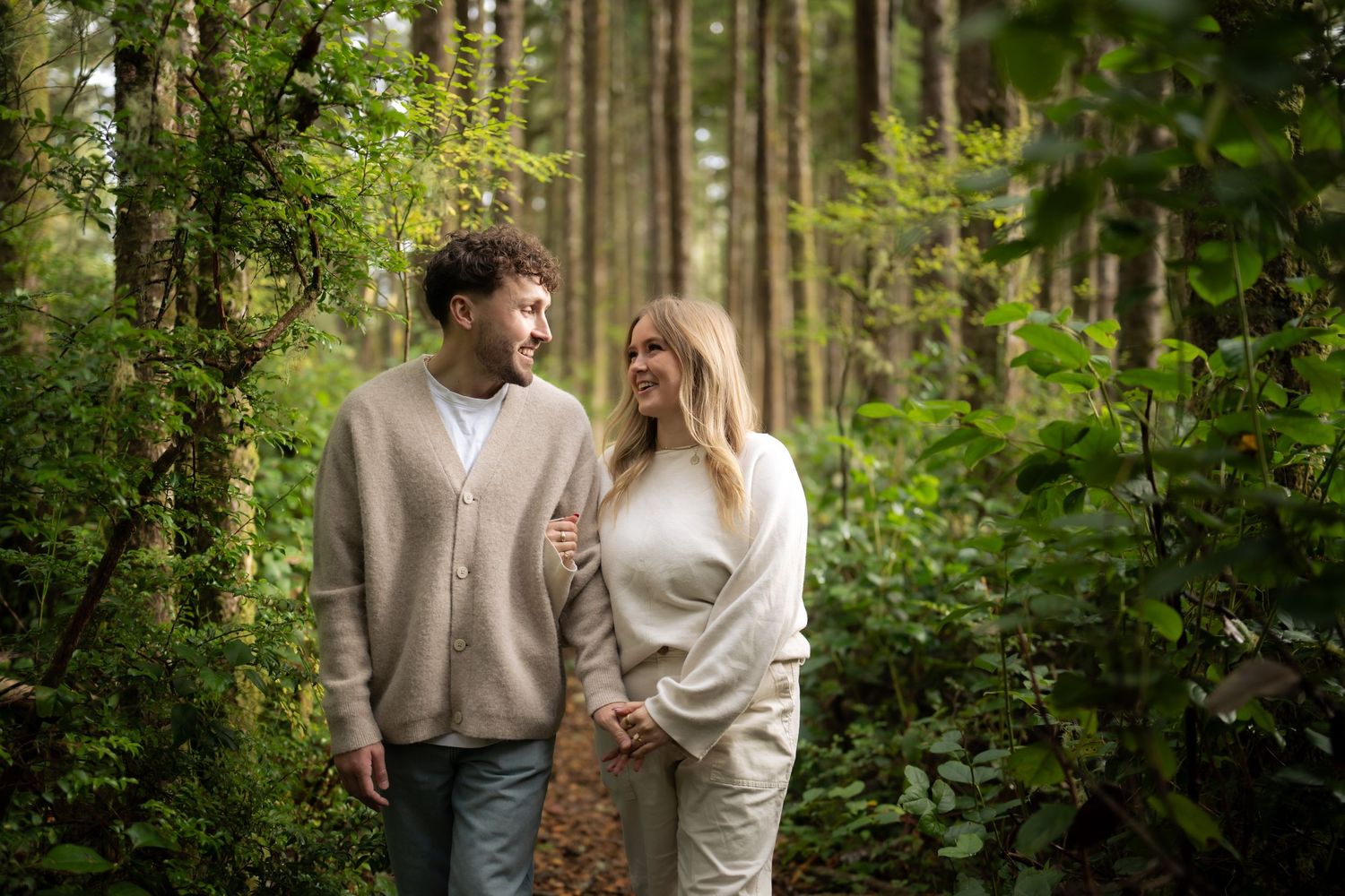Two people stroll intimately through a lush green forest with sunlight filtering through the trees.