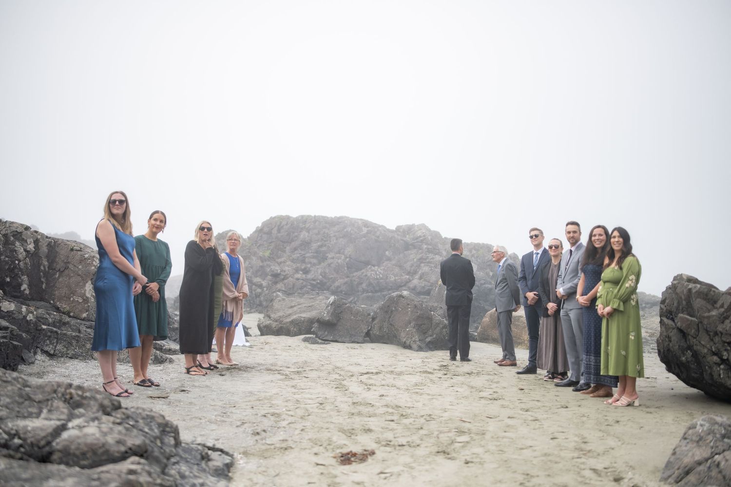 Wedding guests gather on a foggy beach with large rock formations in the background.