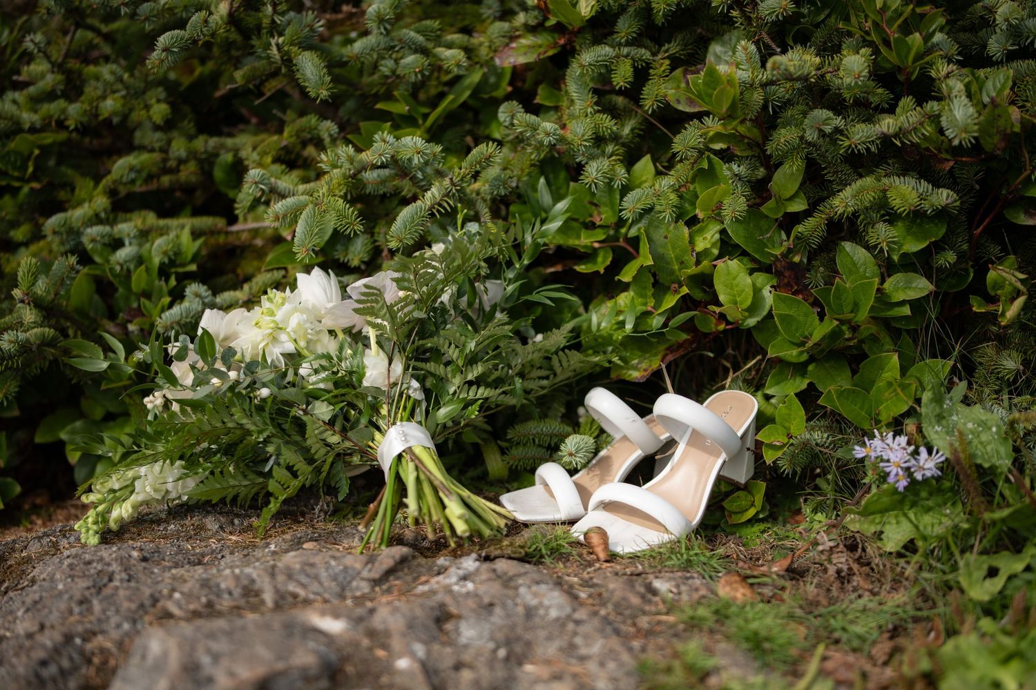 White bridal bouquet and shoes arranged on a rocky surface amid green foliage.