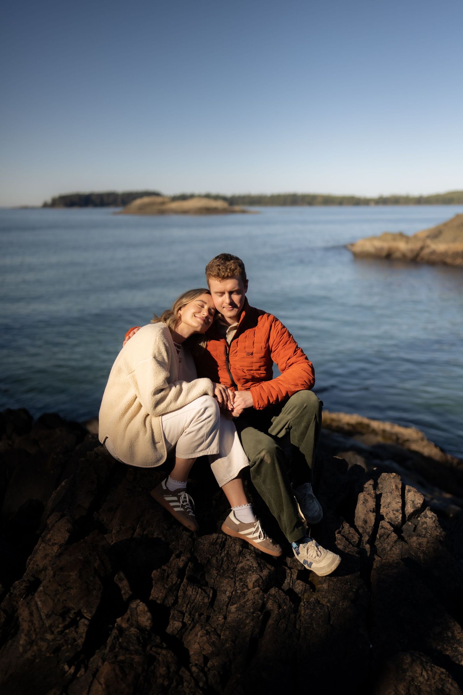 A couple sits close together on dark coastal rocks with ocean views.