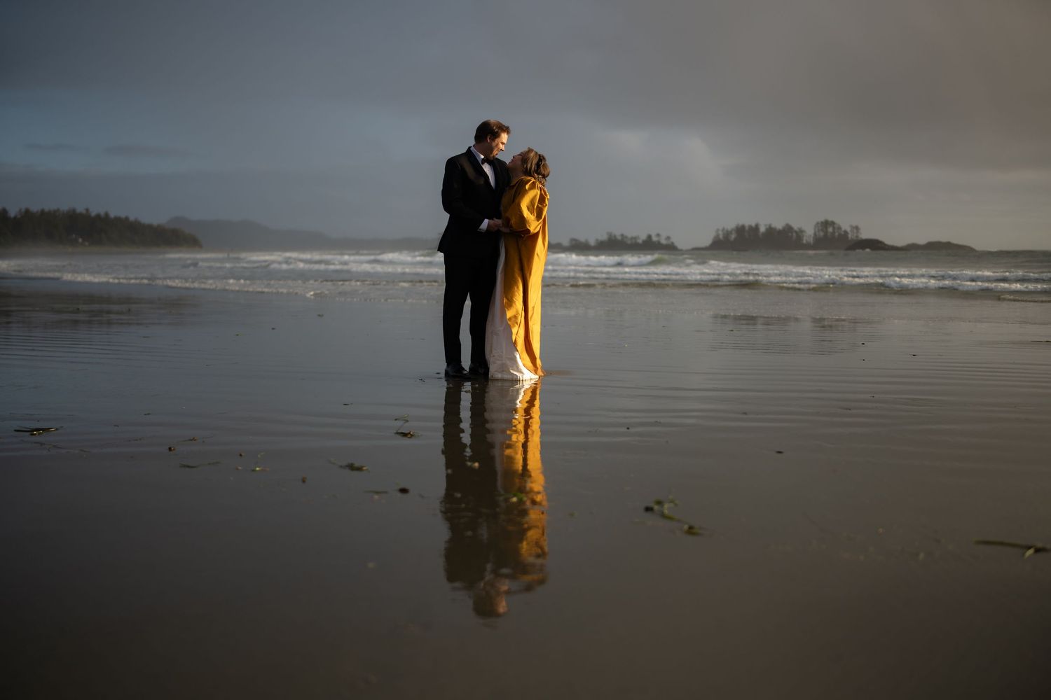 Two people embrace on a misty beach with their reflection visible in the wet sand.