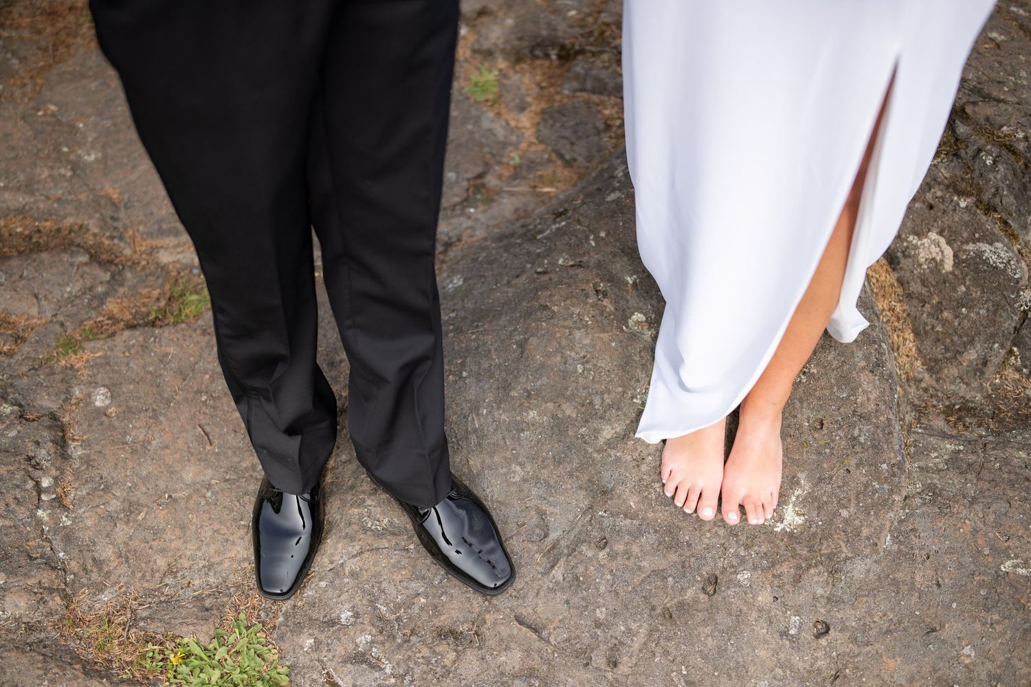 Contrasting formal shoes and bare feet standing on rocky ground outdoors.