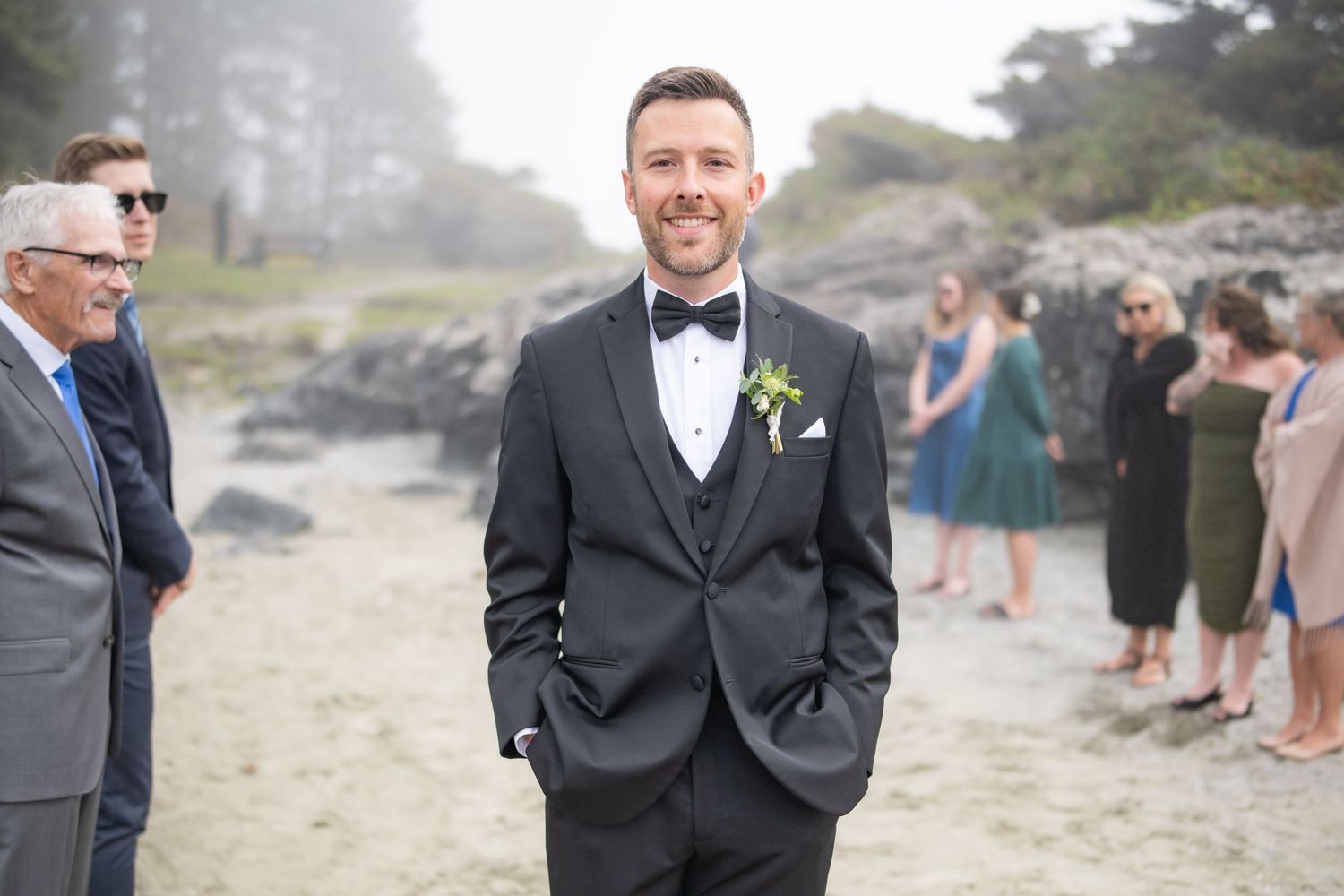 A groom in classic black tuxedo stands on a misty beach with guests in background.
