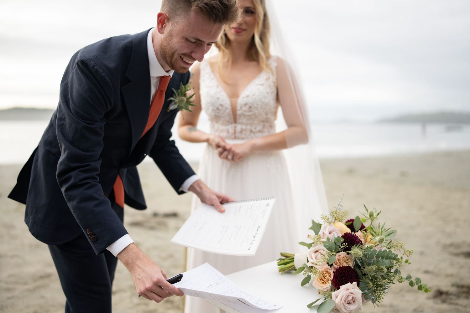 Couple exchanges vows and documents during beach ceremony with floral bouquet.