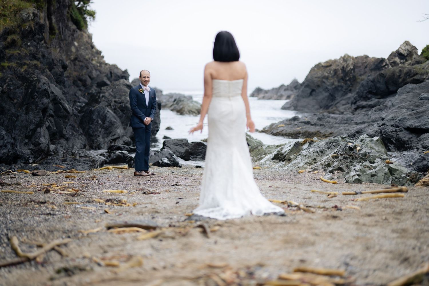 A couple shares an intimate moment on a rocky beach surrounded by evergreen trees and coastal cliffs.