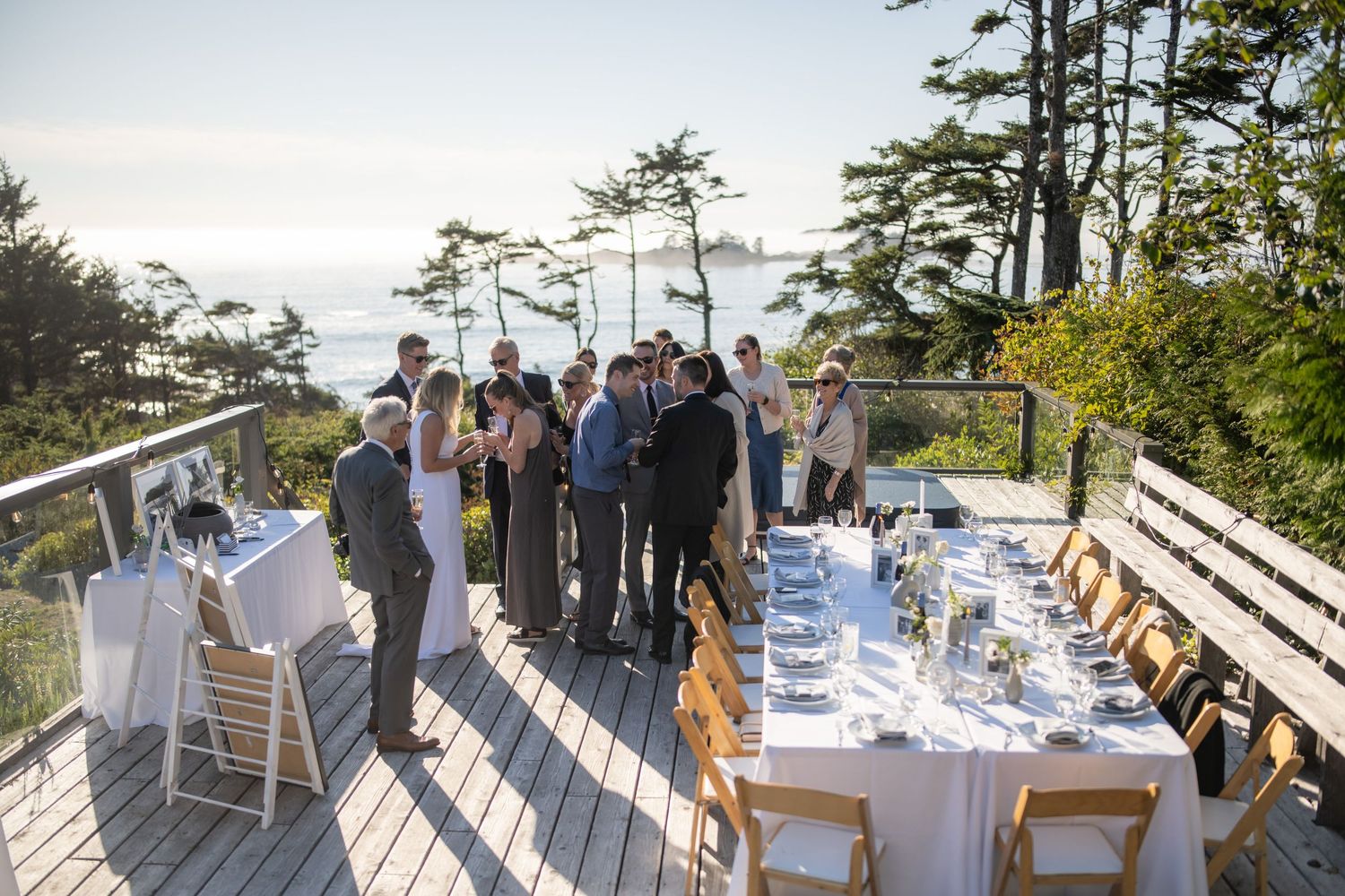 Elegant outdoor reception setup with tables and chairs on a wooden deck overlooking pine trees.