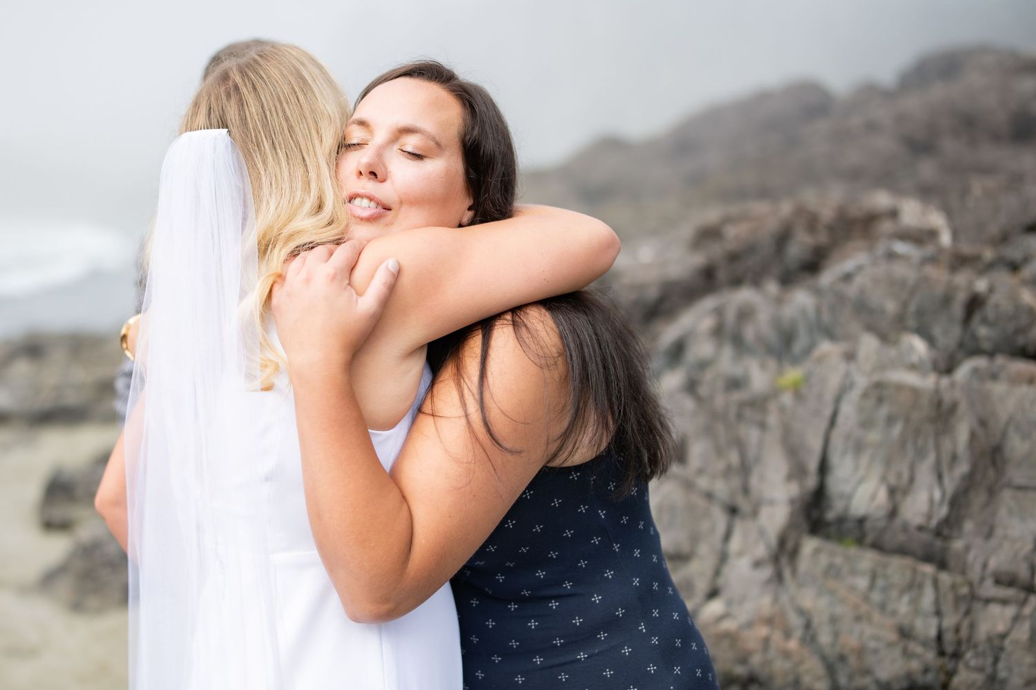 Wedding guest in blue dress hugs bride warmly on rocky beach.