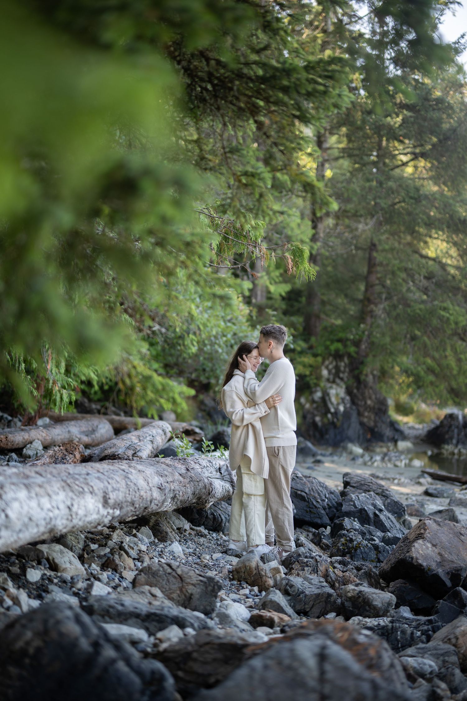 A couple shares an intimate moment on rocky coastal terrain surrounded by evergreen trees.