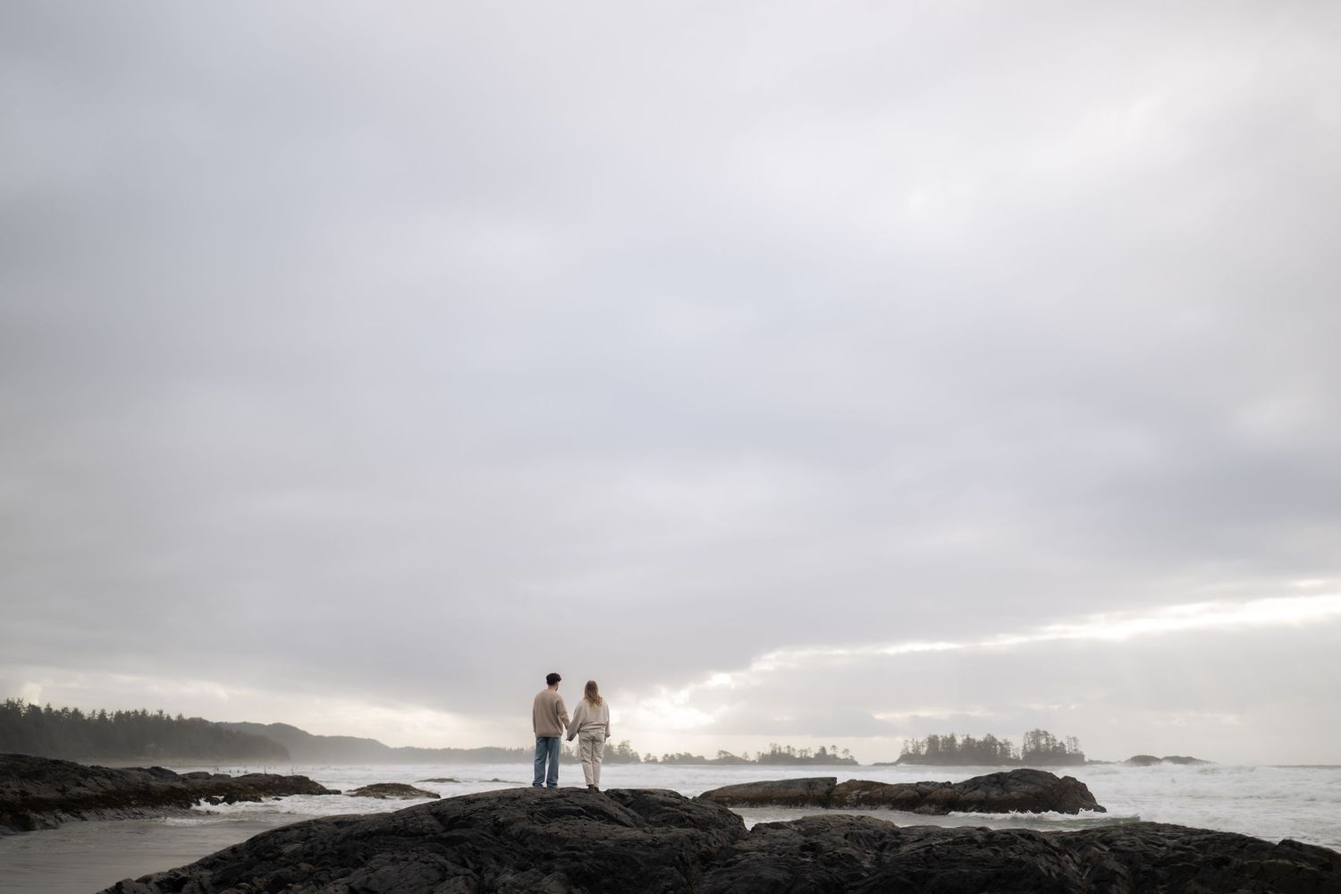 Two figures stand silhouetted against a dramatic cloudy sky on rocky Pacific Northwest coastline.
