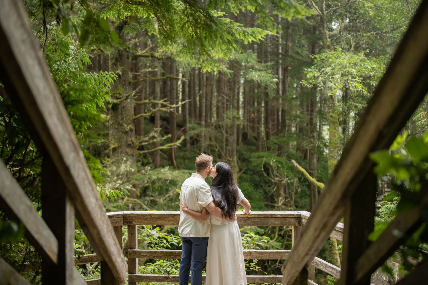 A couple embraces on a wooden bridge surrounded by dense forest greenery.