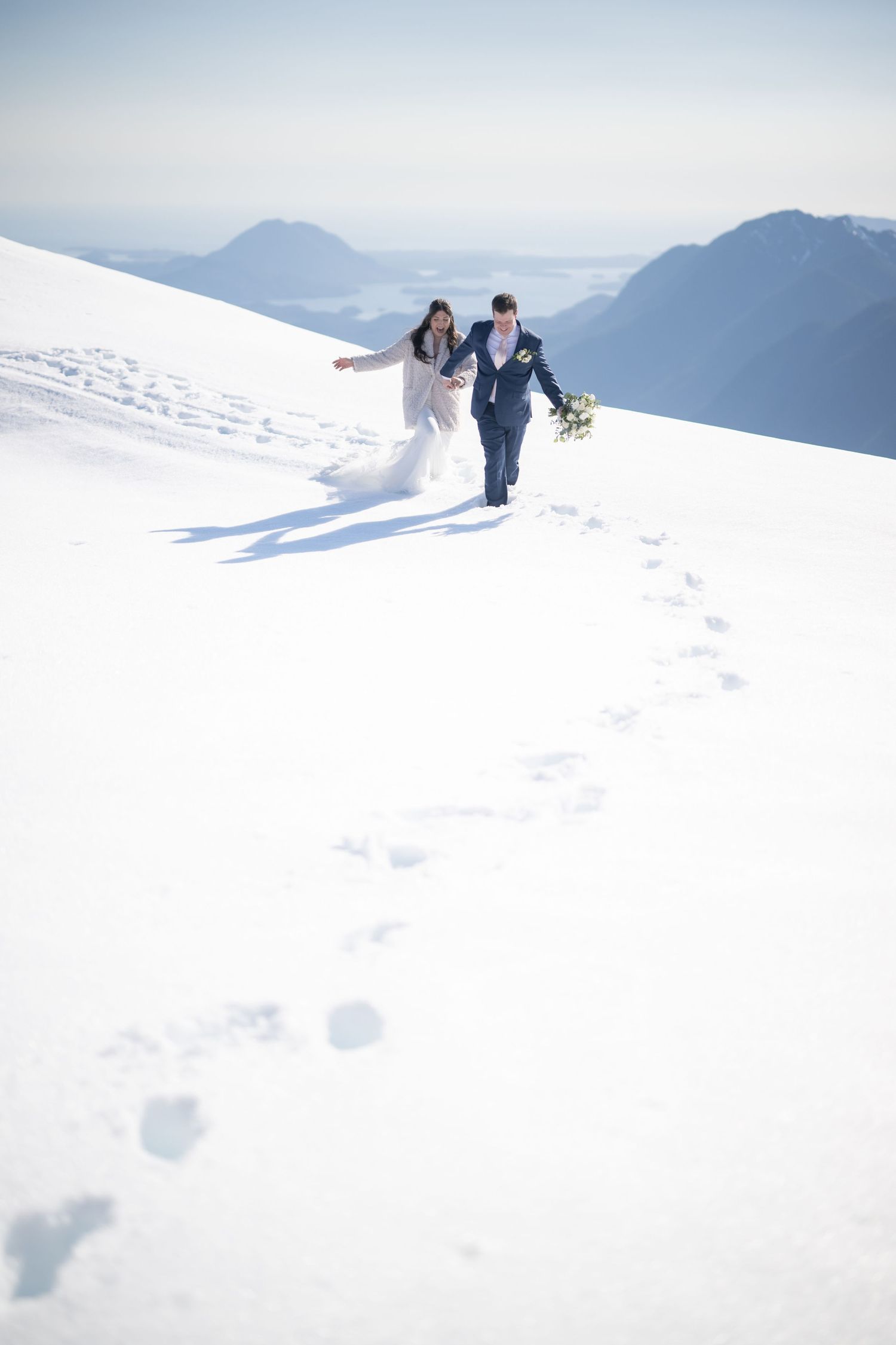 Wedding couple walking through pristine snow with mountain peaks in the distance.