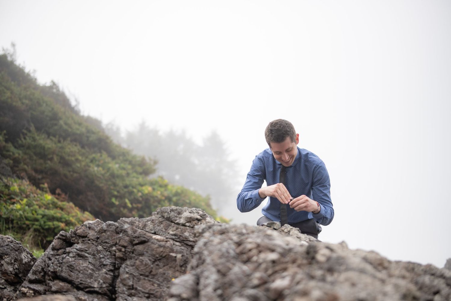 A person in a blue jacket crouches on a misty rocky outcrop.