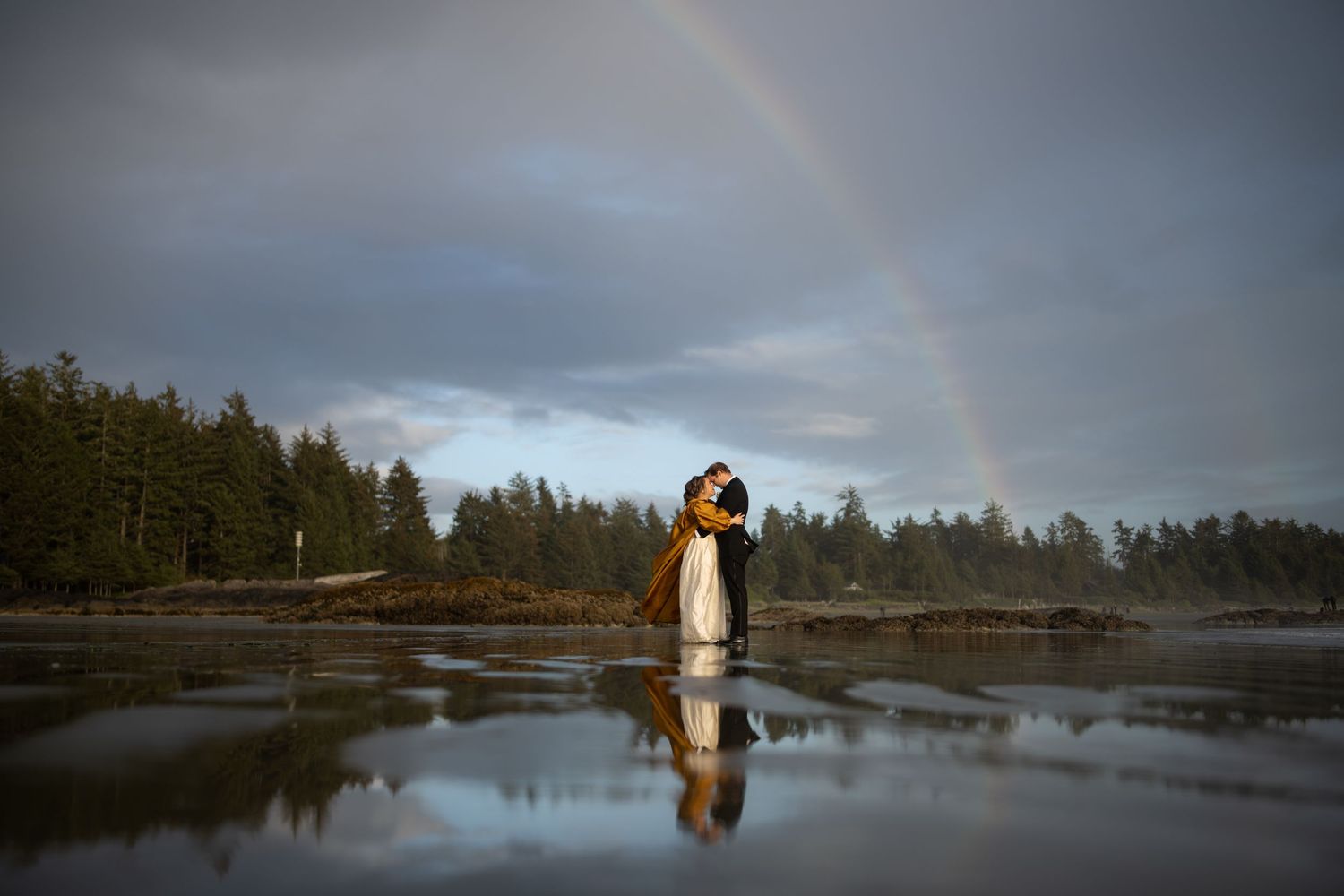 A beautiful rainbow arches over a couple standing in reflection on a wet beach.