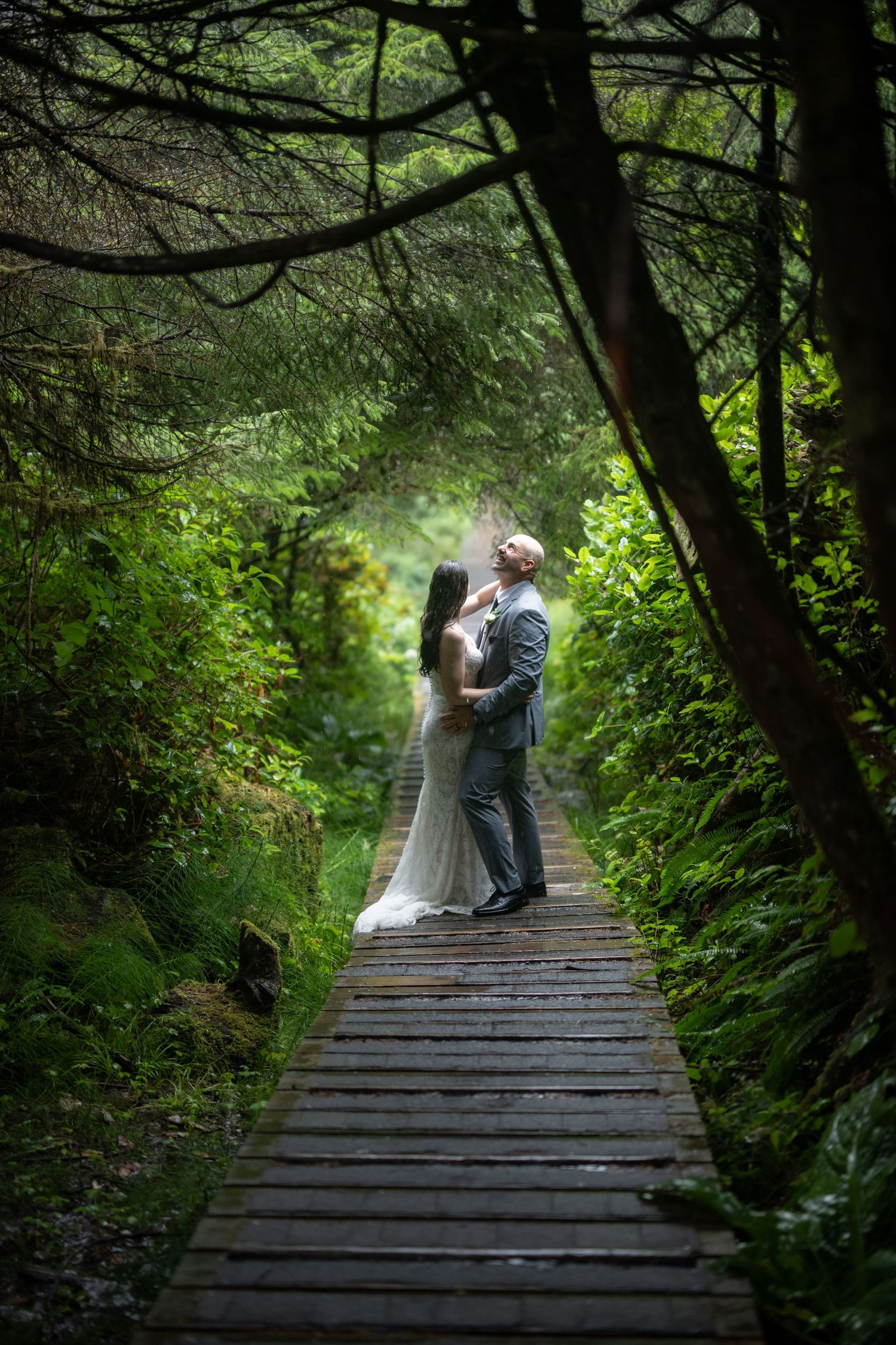 Couple stands together on wooden boardwalk surrounded by dense forest vegetation.