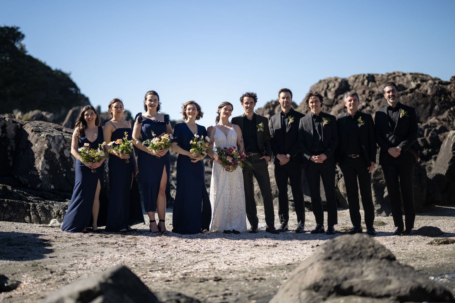 Wedding group photo with bridal party lined up against dramatic coastal backdrop.
