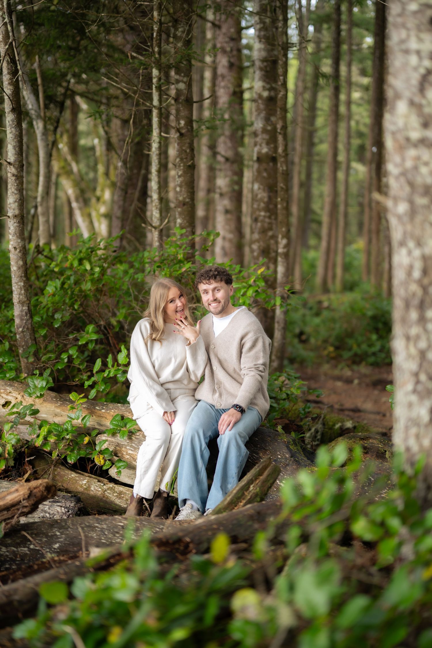 Two people share an intimate moment while seated on a log in a lush evergreen forest.