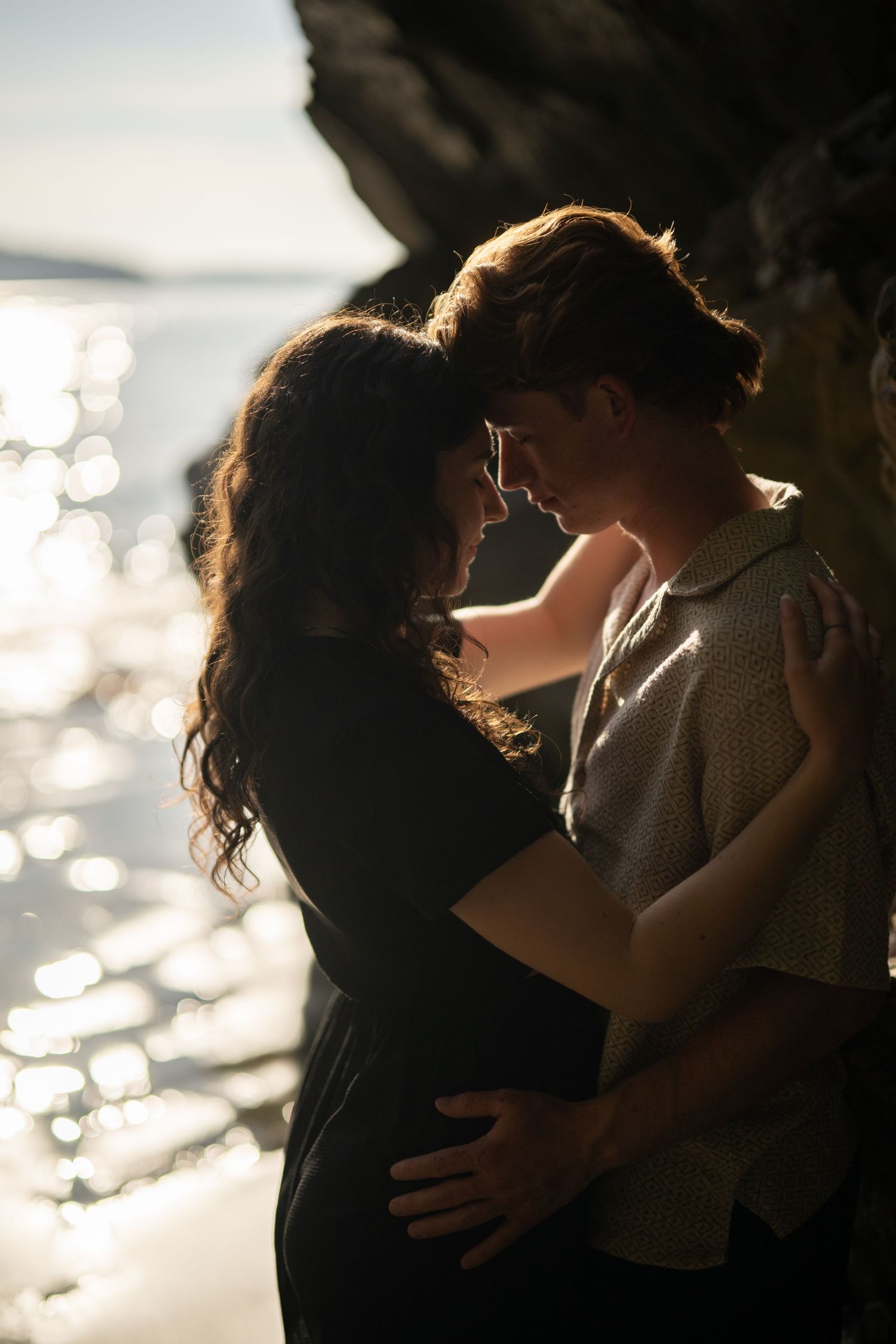 Backlit silhouettes embrace near coastal rocks with sparkling water in background.