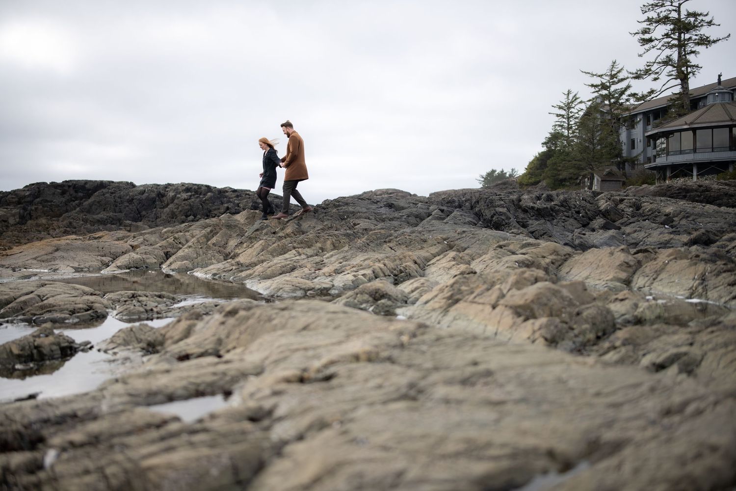 Two figures walk along rocky coastal cliffs with a house visible in the background.
