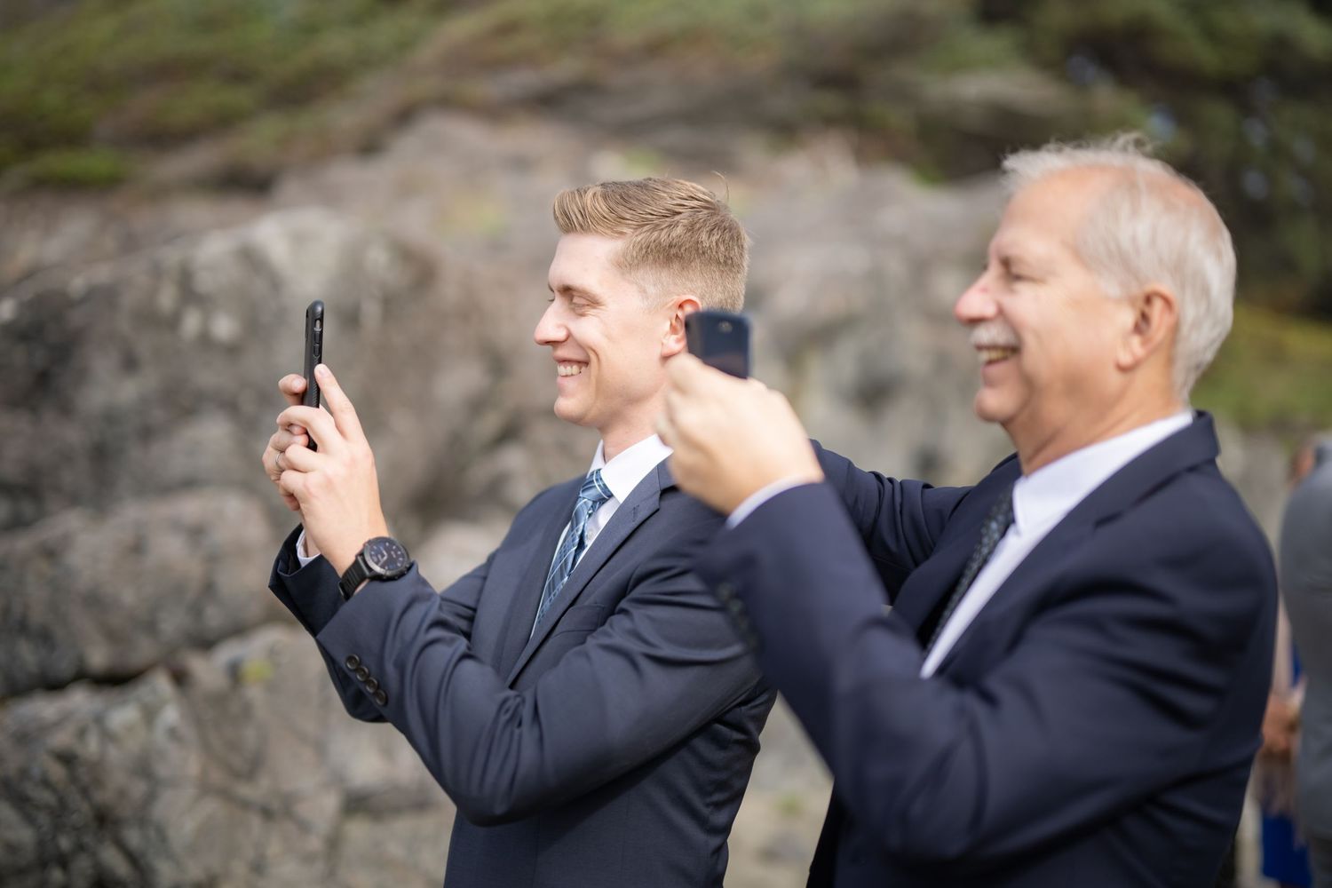 Wedding guests taking photos with phones on rocky cliff.