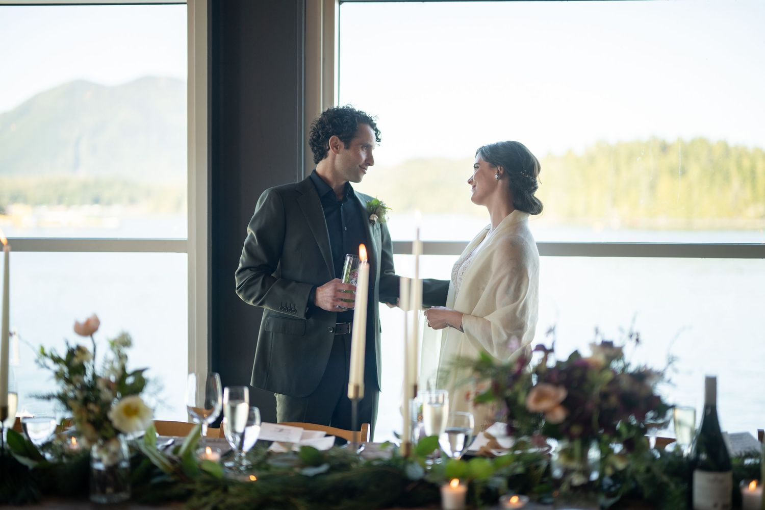 Two people sharing drinks by large window with mountain and water views.