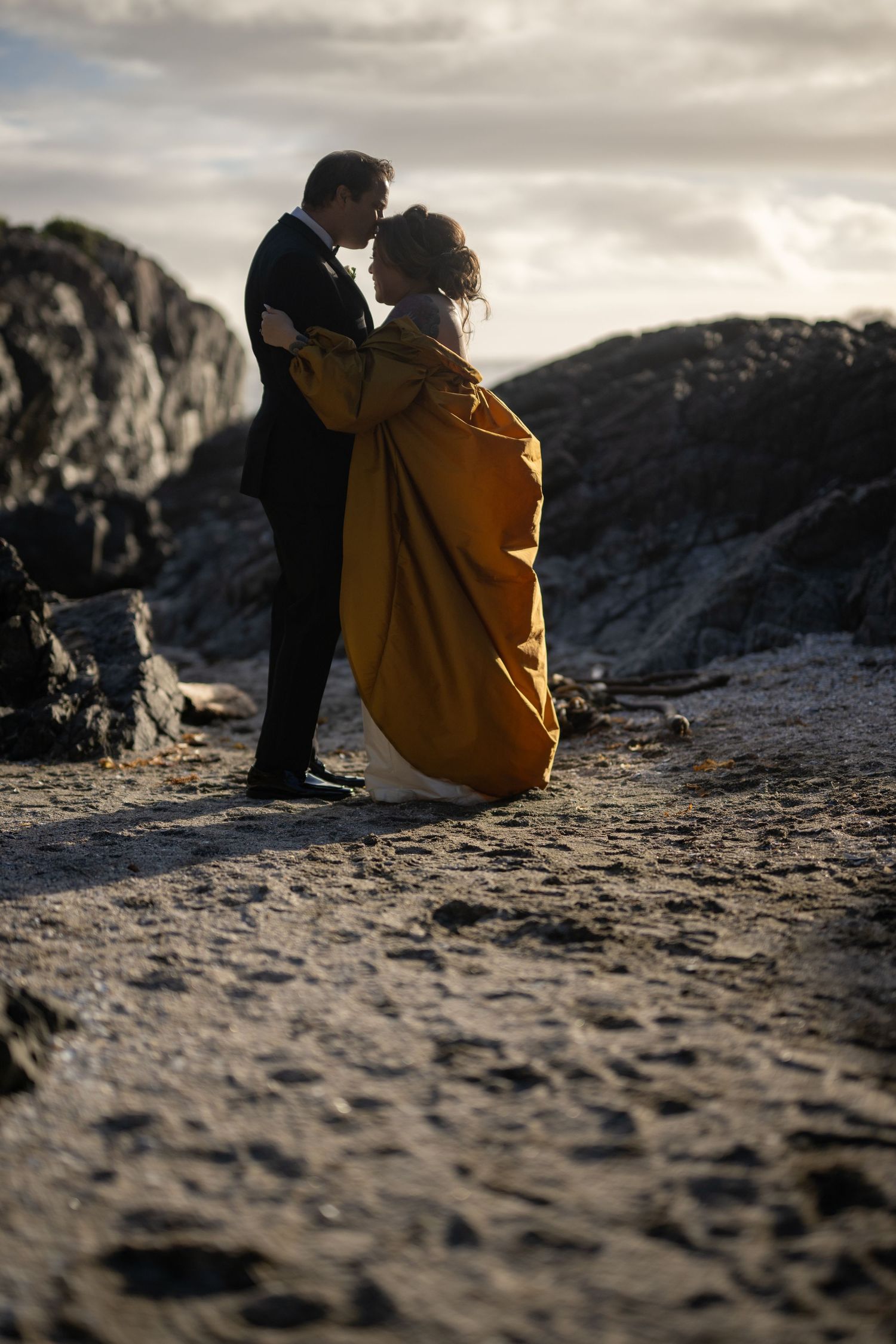 Two people sit together on a rocky beach during sunset.